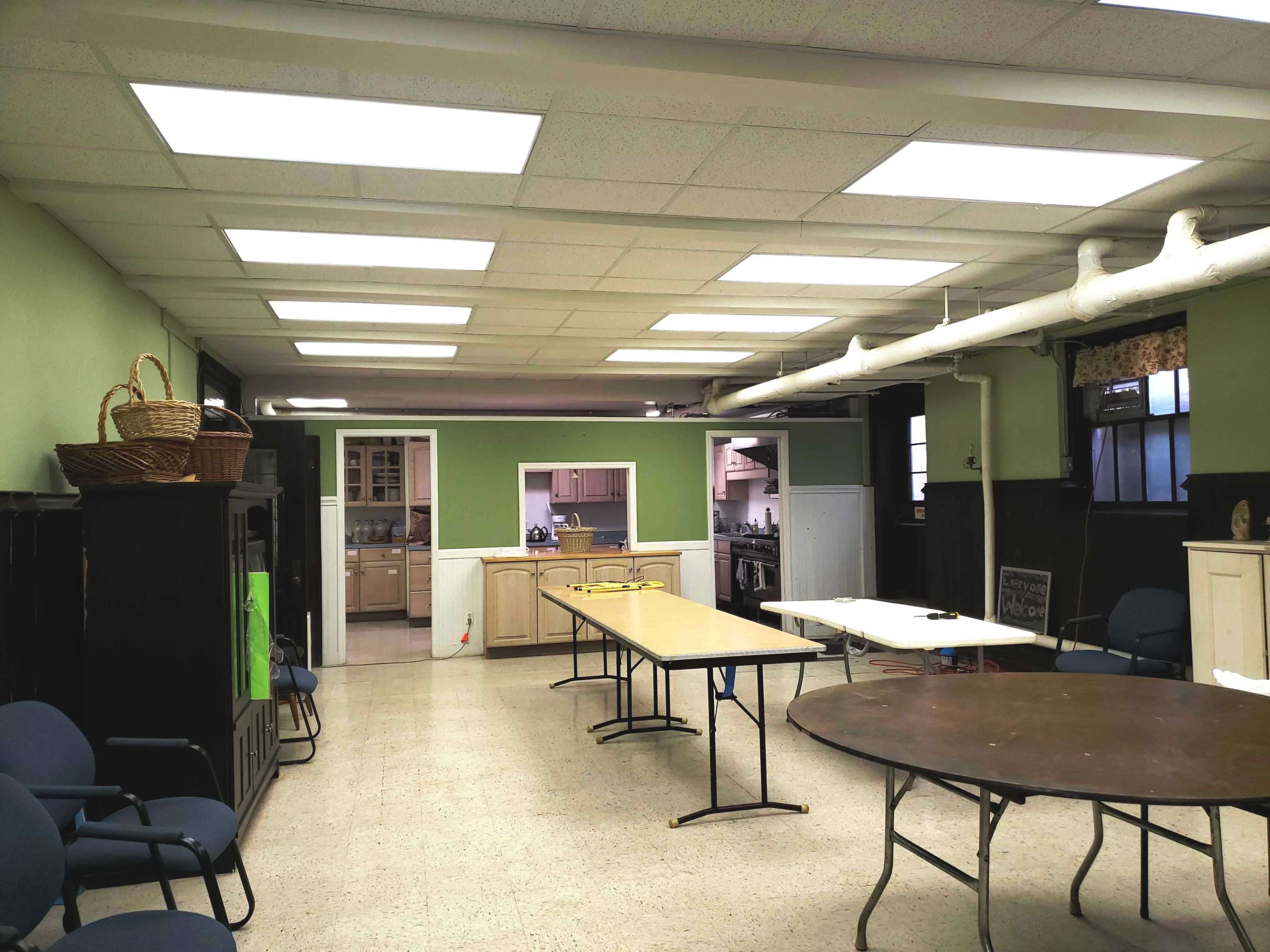 An empty room with several tables arranged, light-colored walls, and a kitchen area visible in the background.