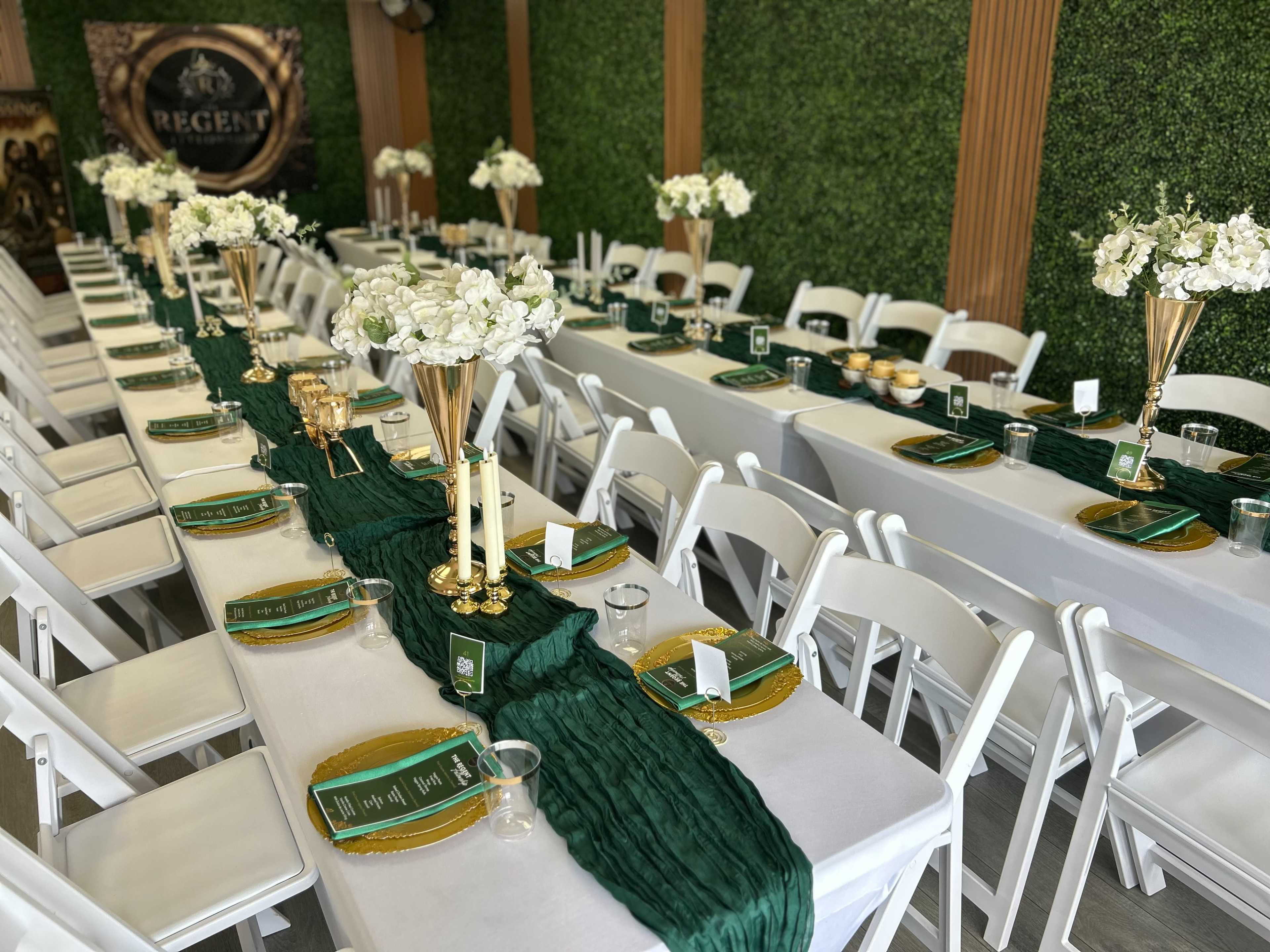 The image shows a dining setup with long tables adorned with green fabric runners, white floral centerpieces, and golden decorative elements in a venue with a green wall backdrop.