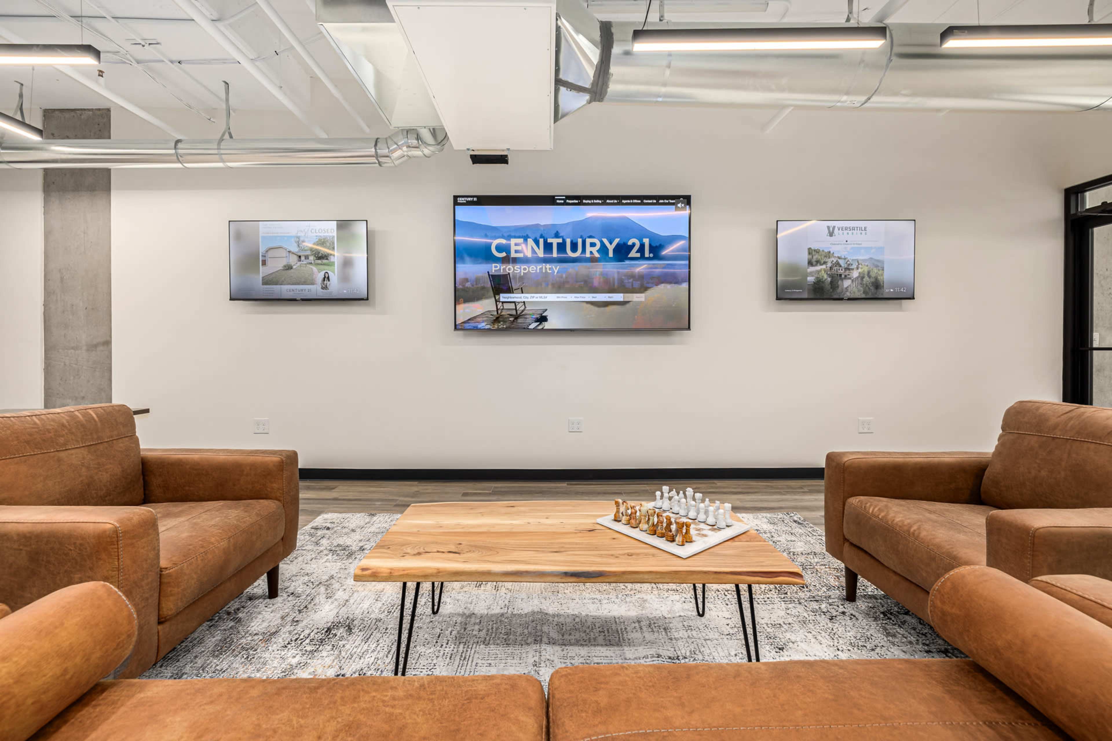 The image shows a modern office lounge area with brown leather sofas, a wooden coffee table featuring a chess set, and three screens displaying the "CENTURY 21" logo and images.