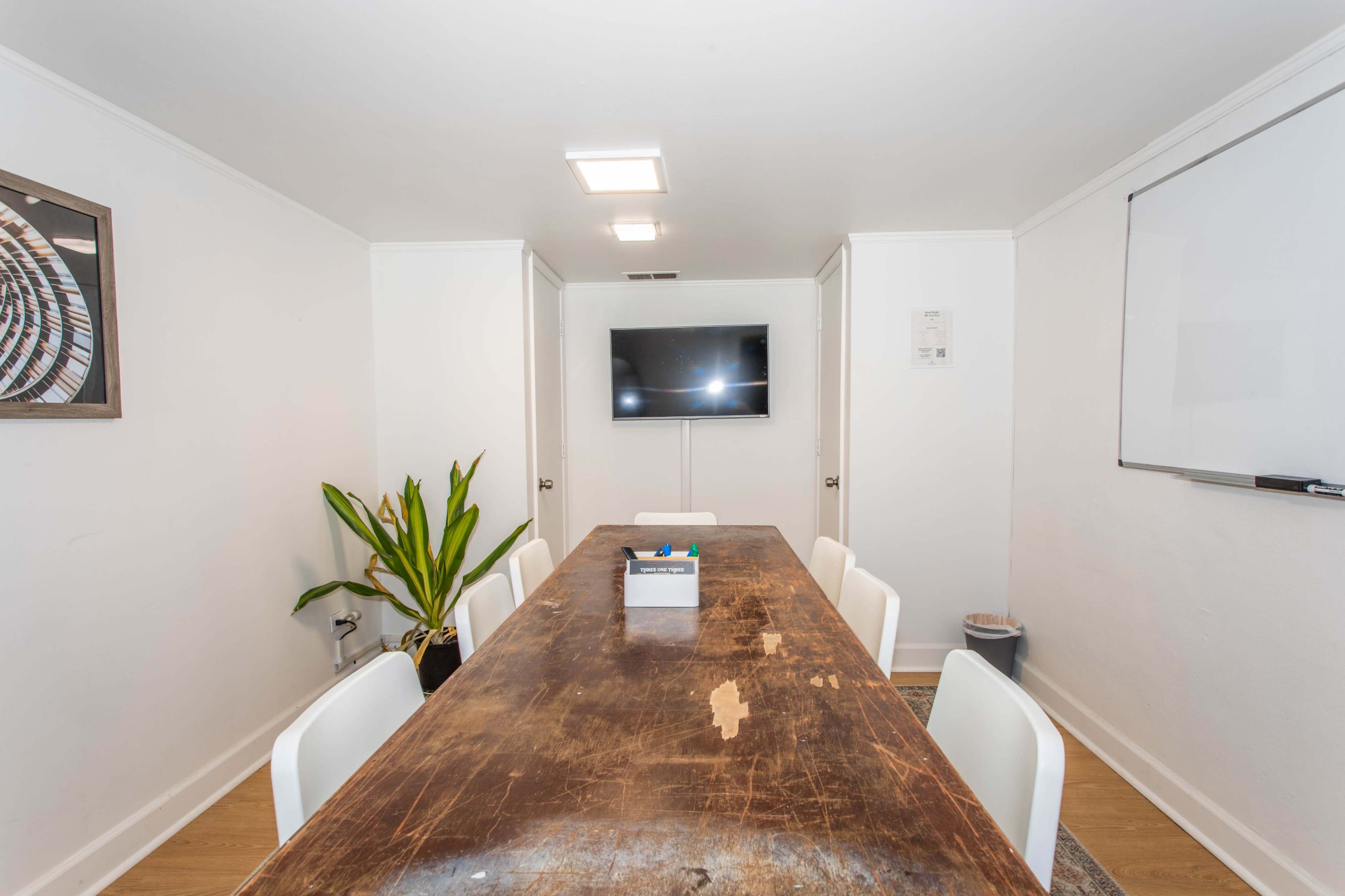 A long, wooden table is surrounded by white chairs in a well-lit meeting room featuring a television and a whiteboard on the walls.