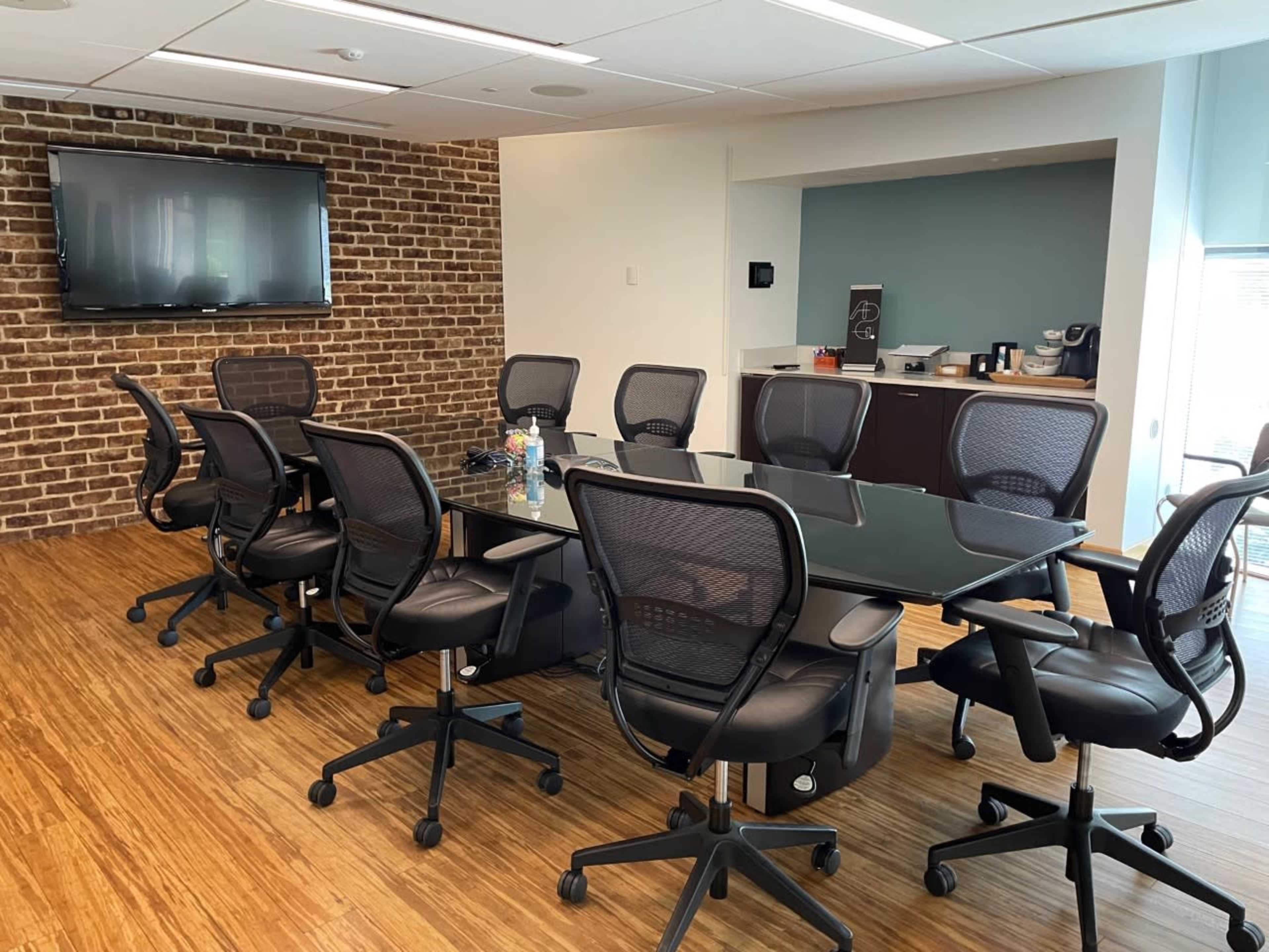 A conference room features a large glass table surrounded by black mesh chairs, with a brick wall and a television mounted on one side.