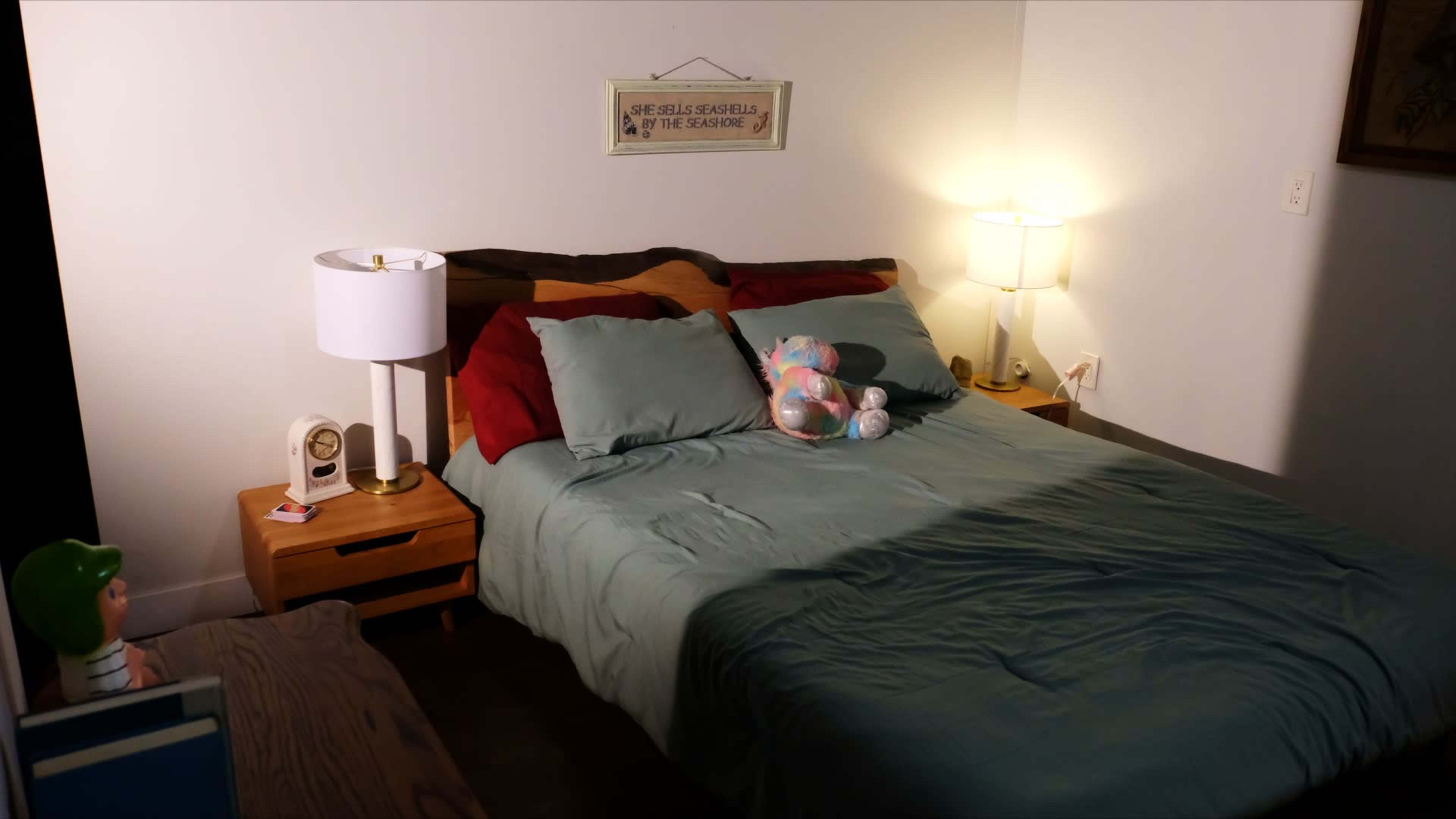 A neatly made bed with teal sheets and decorative pillows is positioned between two bedside tables, each holding a lamp and personal items, in a softly lit bedroom.