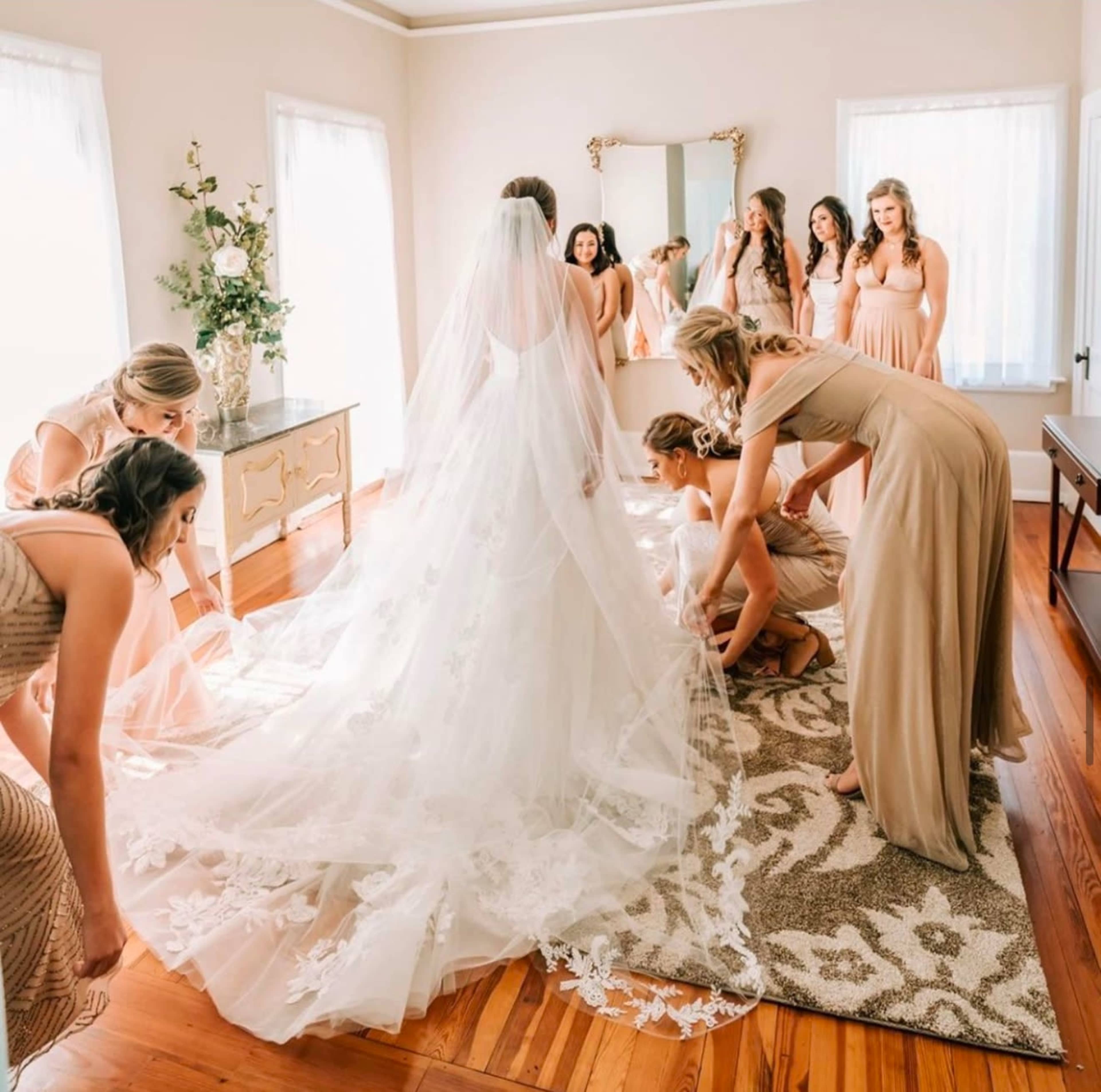 A bride in a wedding dress stands in front of a mirror while her bridesmaids adjust her train in a well-lit room.