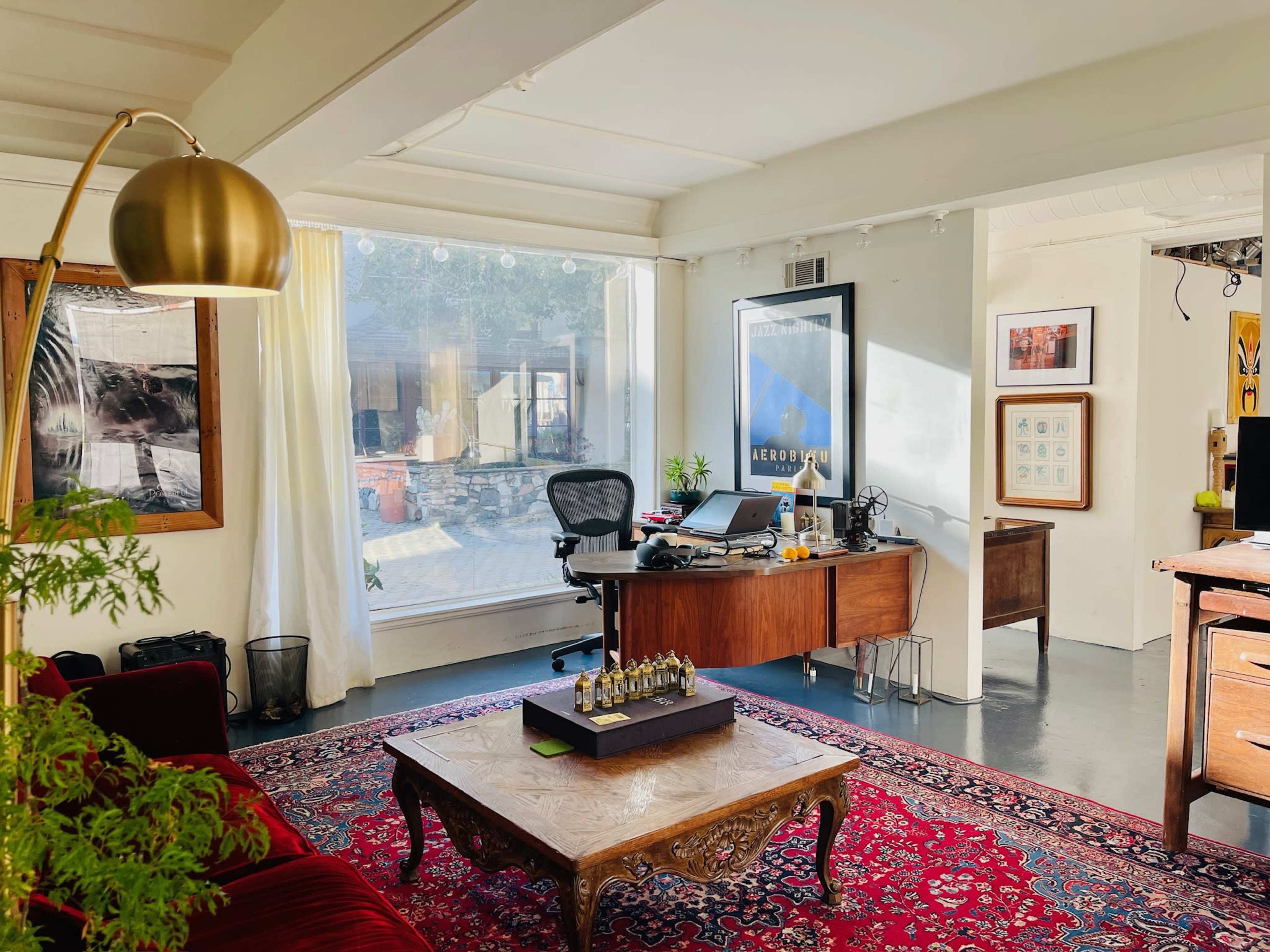 The image shows a modern office space with a wooden desk, a comfortable chair, and a decorative area rug, all illuminated by natural light from large windows.