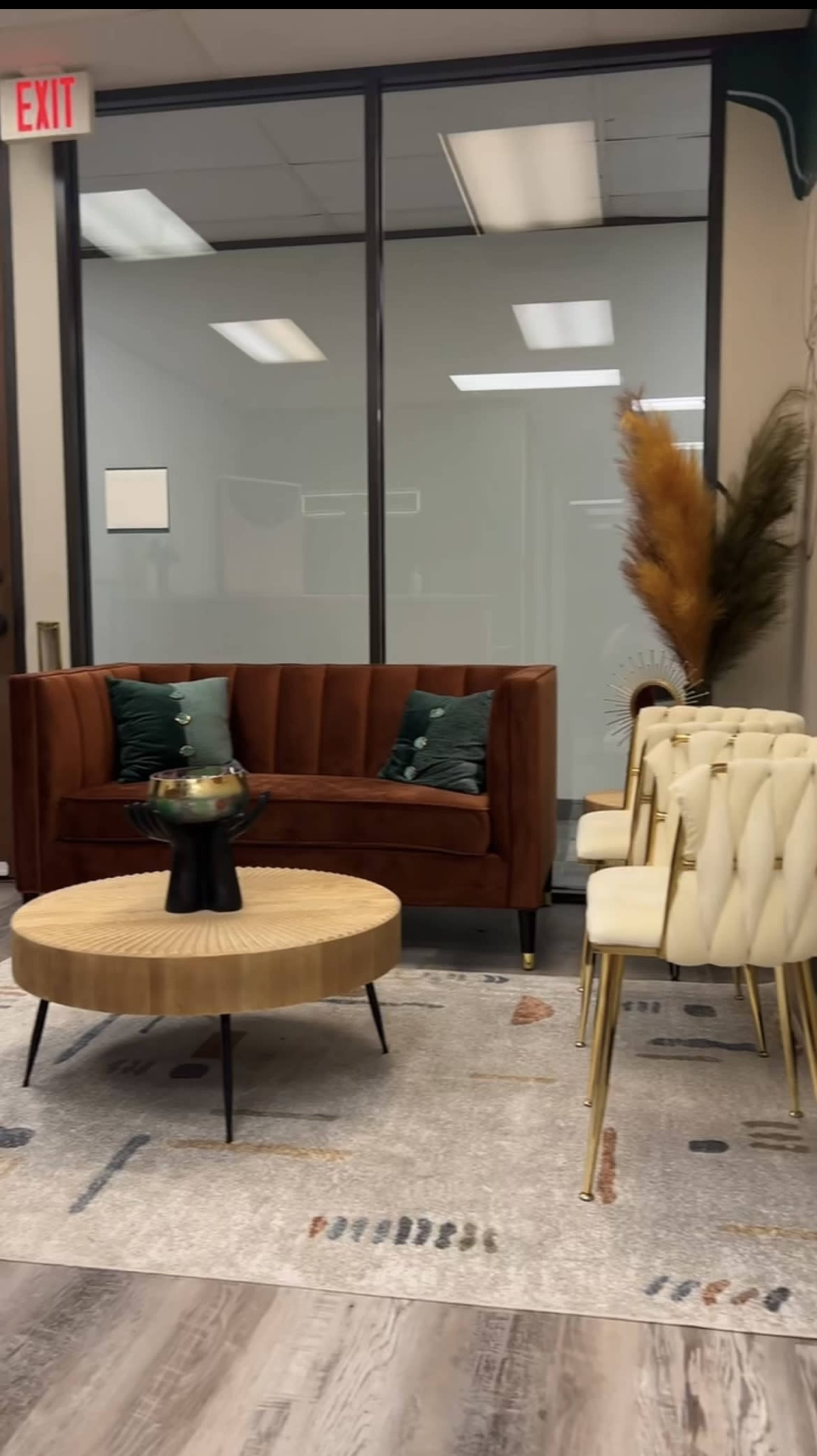 A modern office waiting area features a brown velvet sofa with green cushions, a round wooden coffee table, white upholstered chairs, and decorative plants.