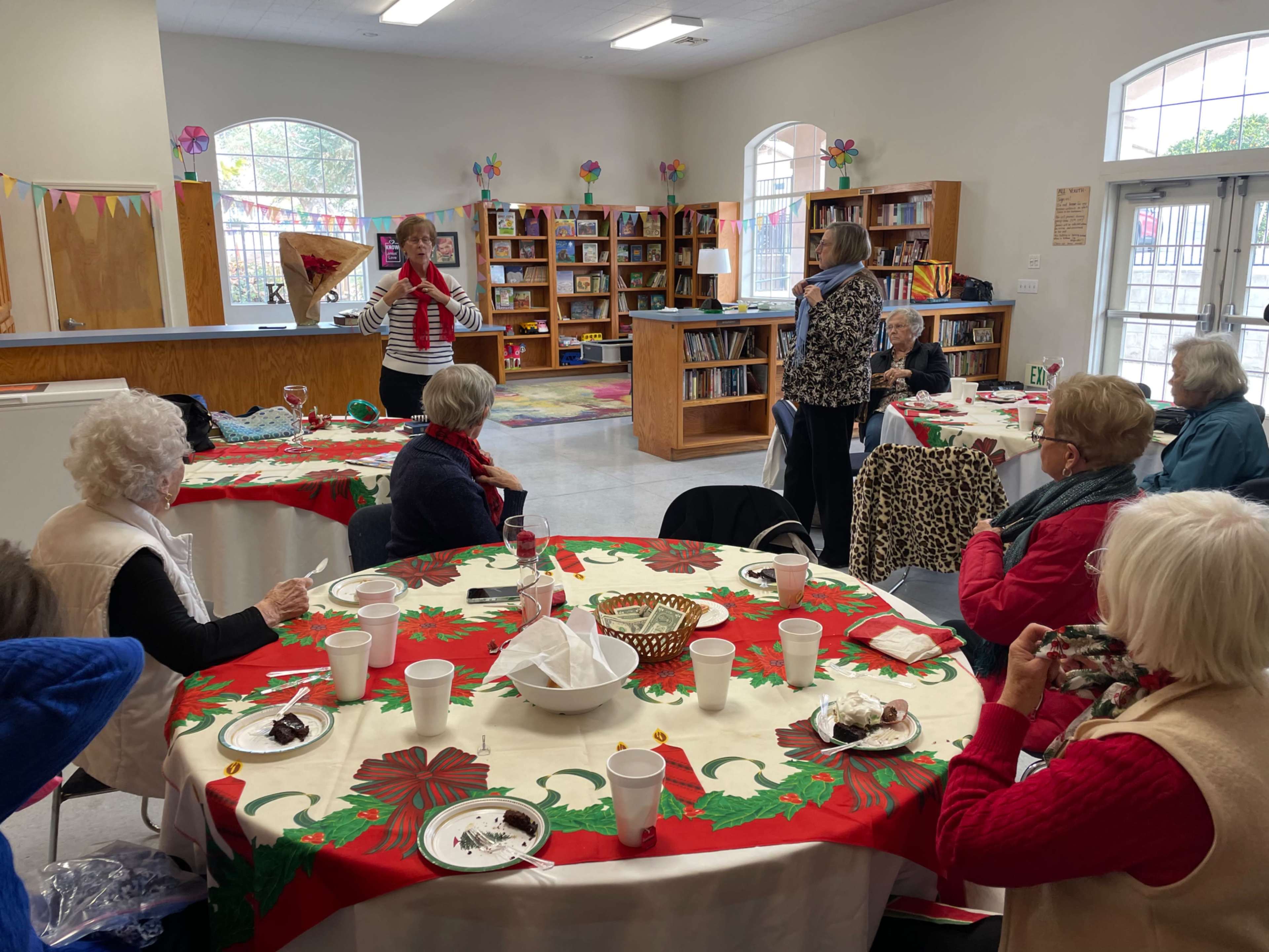 A group of seniors gathers around tables decorated for a festive event in a community room with bookshelves and colorful decorations.