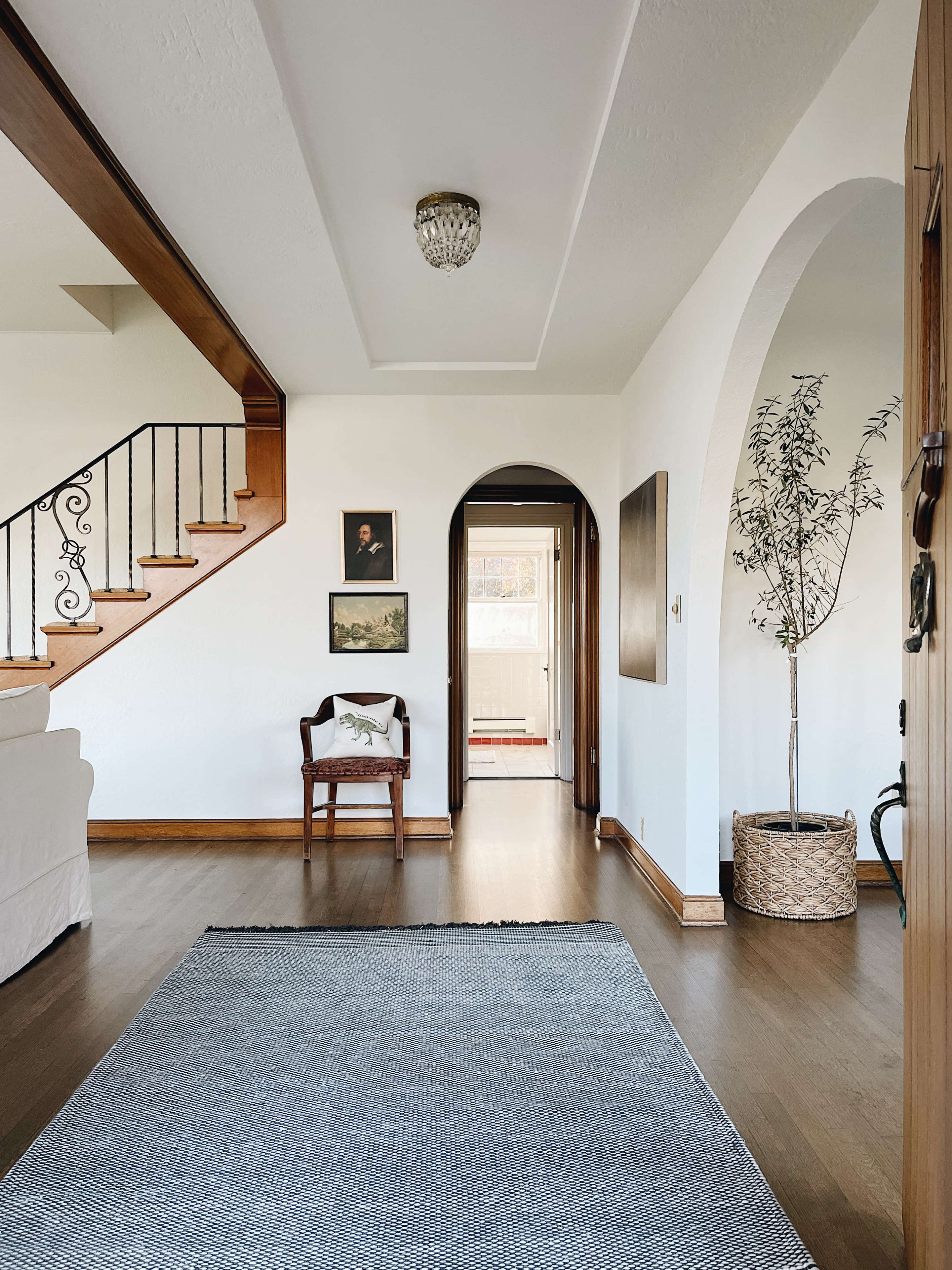 A hallway features a staircase with decorative railing, an arched doorway, framed art on the walls, and a chair beside a potted plant.