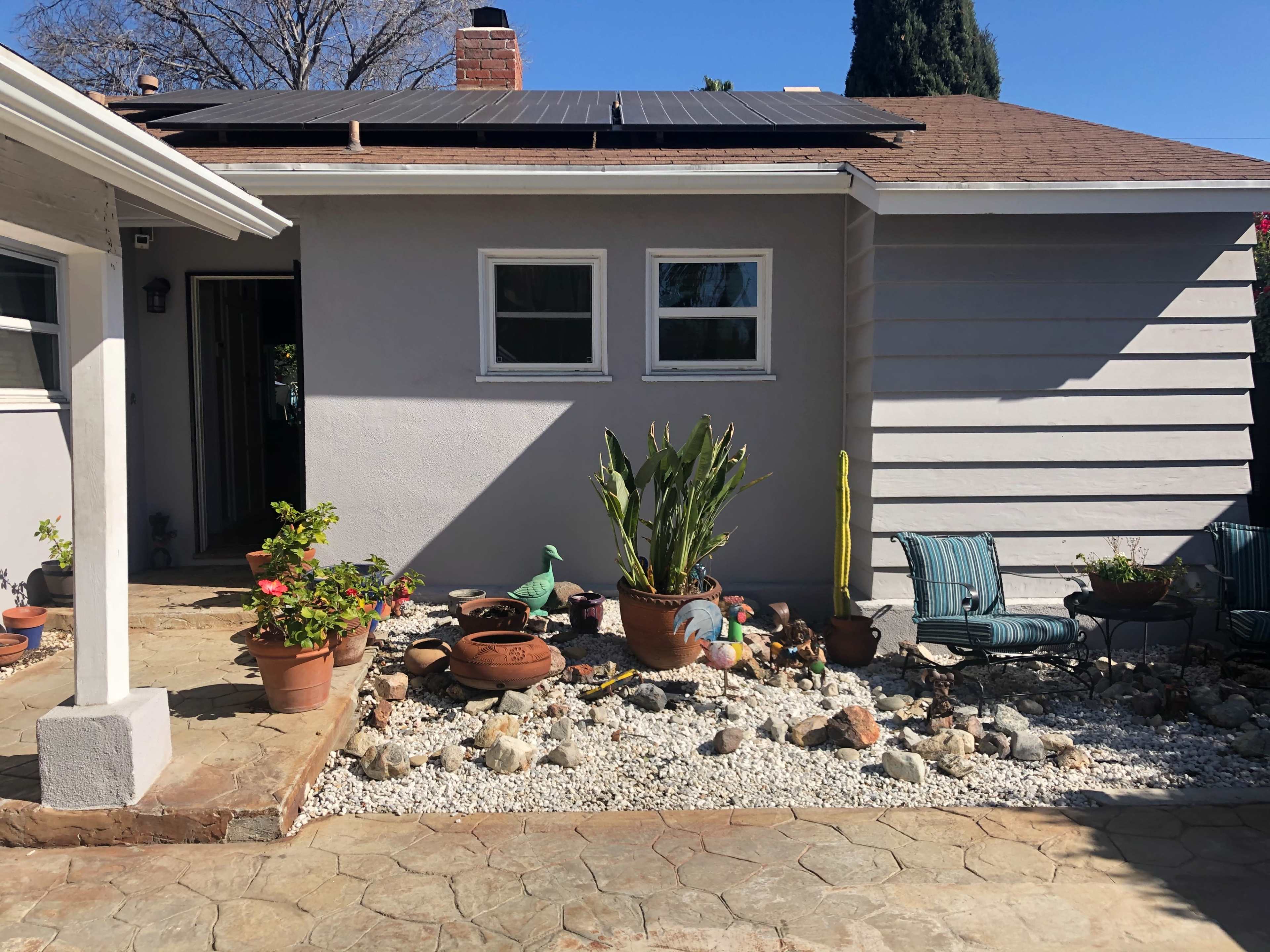 A house facade with solar panels on the roof, surrounded by a garden featuring potted plants and gravel.