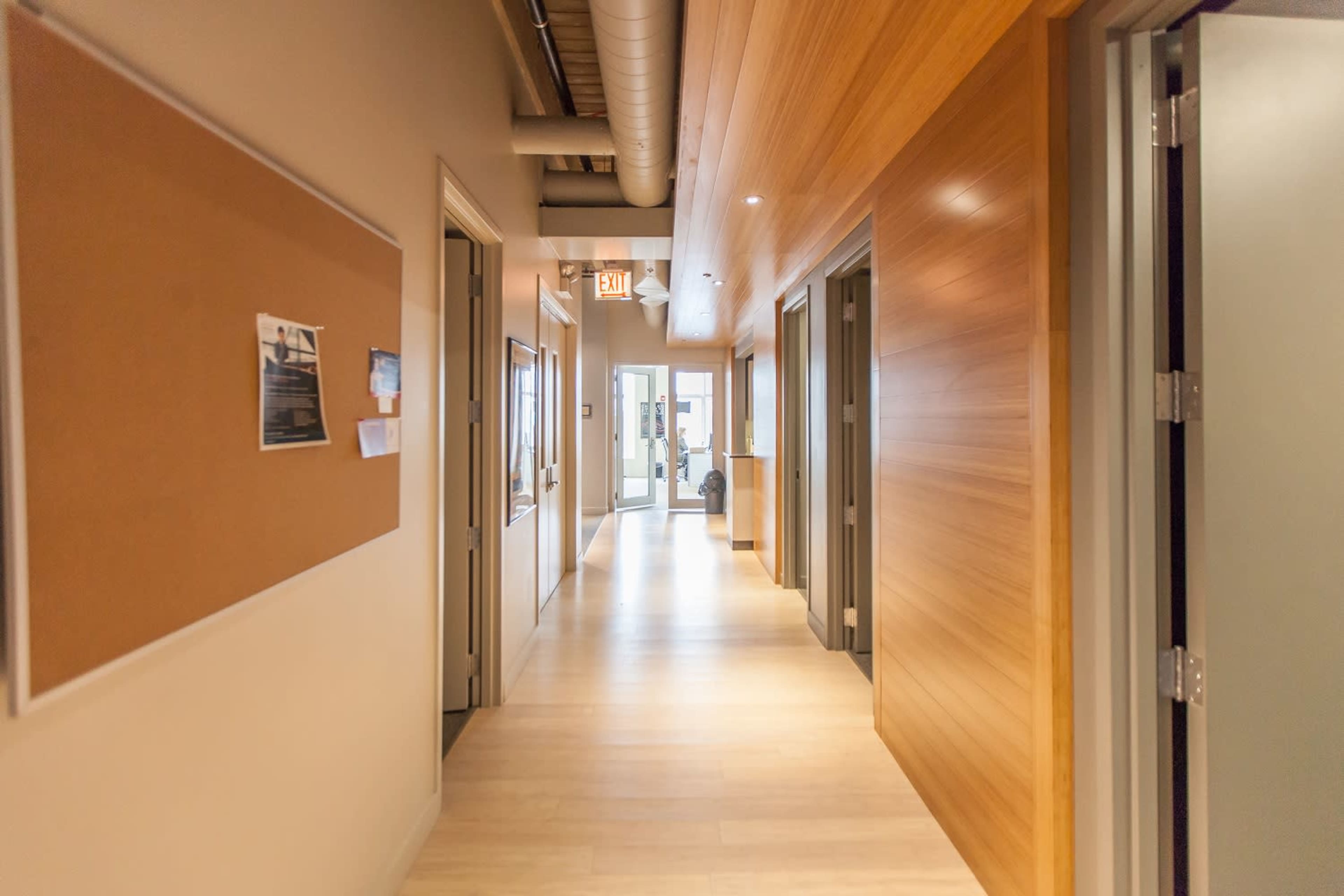 A wide corridor with wooden walls and several doorways leading to offices or rooms, ending with a glass door at the far end.