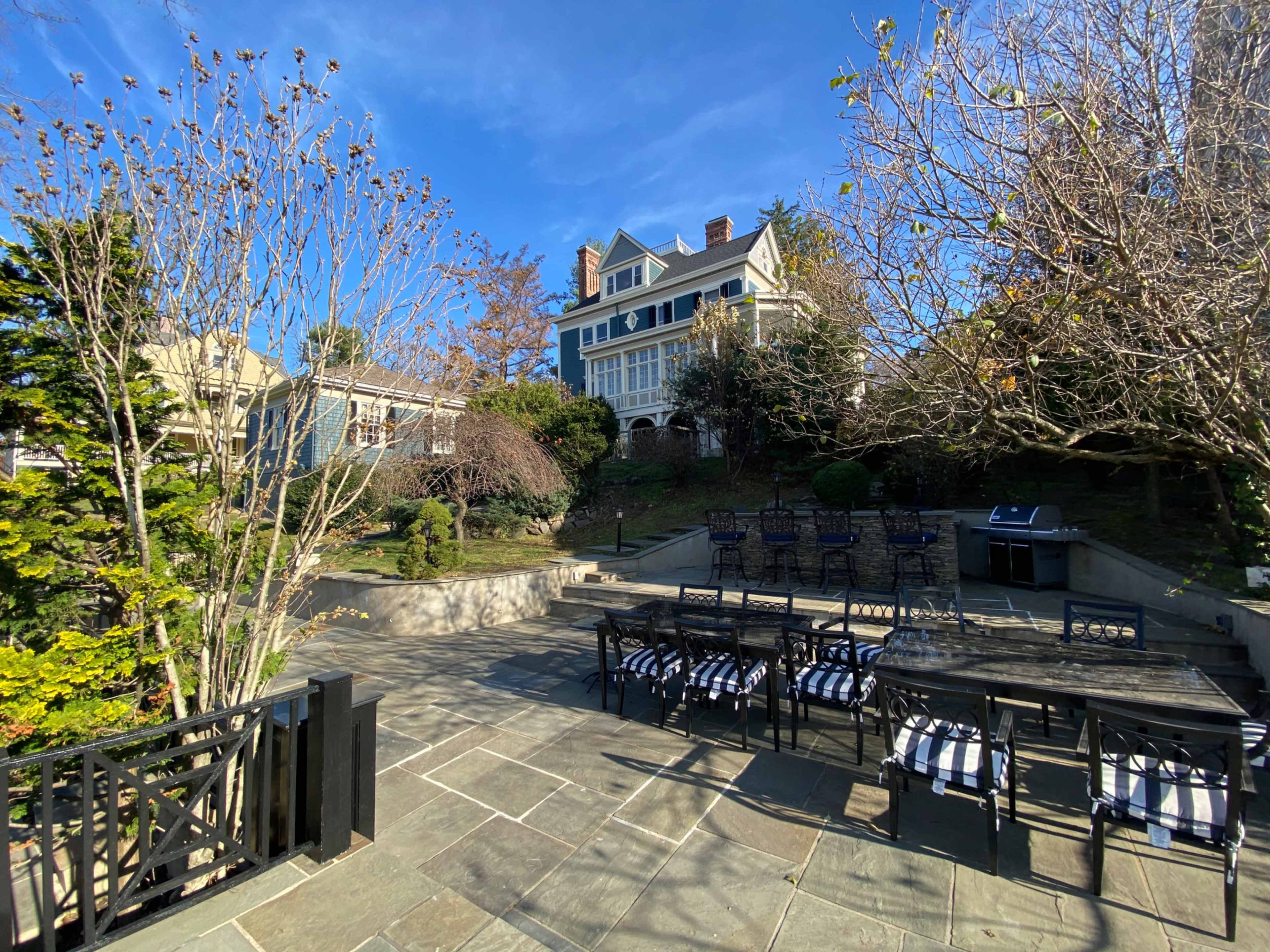 A large stone patio with outdoor seating and dining furniture is situated in front of a multi-story house surrounded by trees and shrubs.