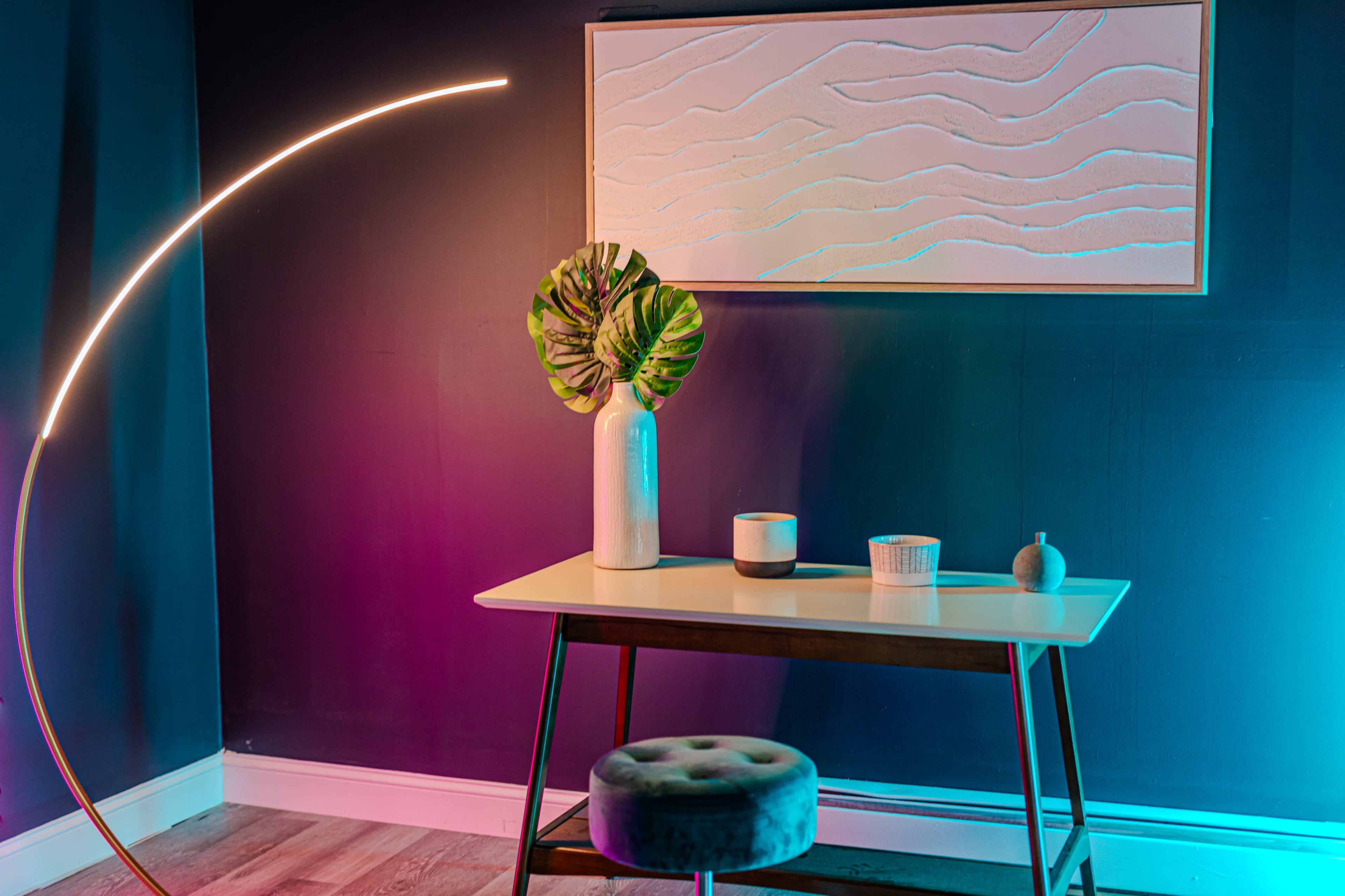 A small table with a vase and decorative items is set against a dark blue wall, illuminated by colorful lighting and a curved floor lamp.