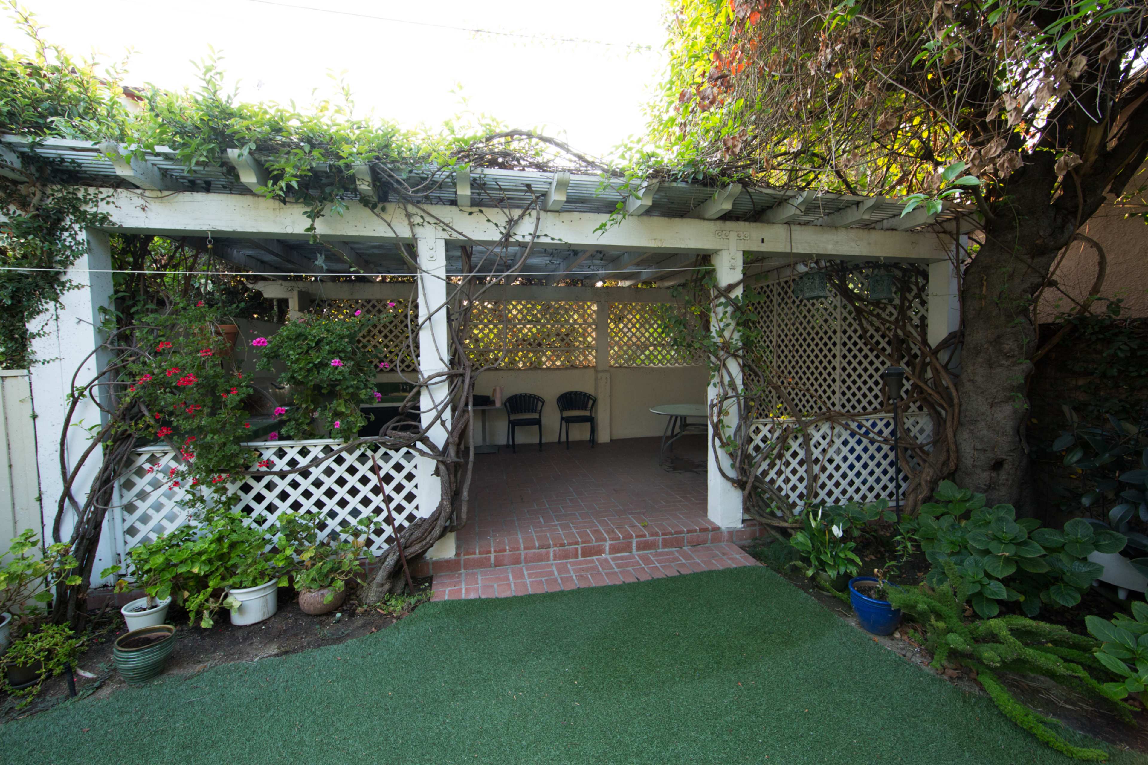 A wooden pergola covered with vines and flowers frames a small patio area with chairs, surrounded by greenery and potted plants.