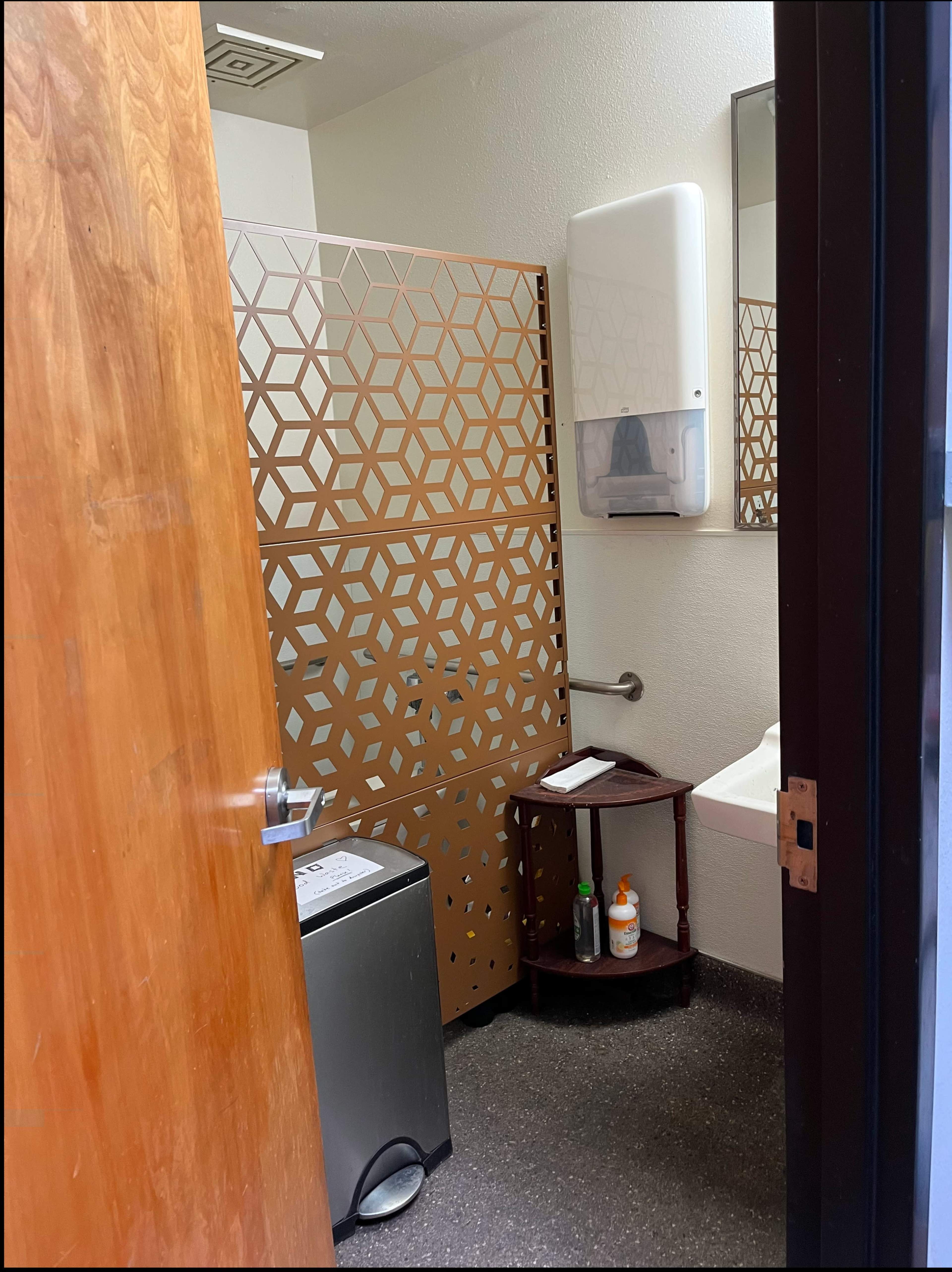 The image shows a partially open restroom door revealing a modern bathroom with a decorative partition, a sink, and various dispensers on the wall.