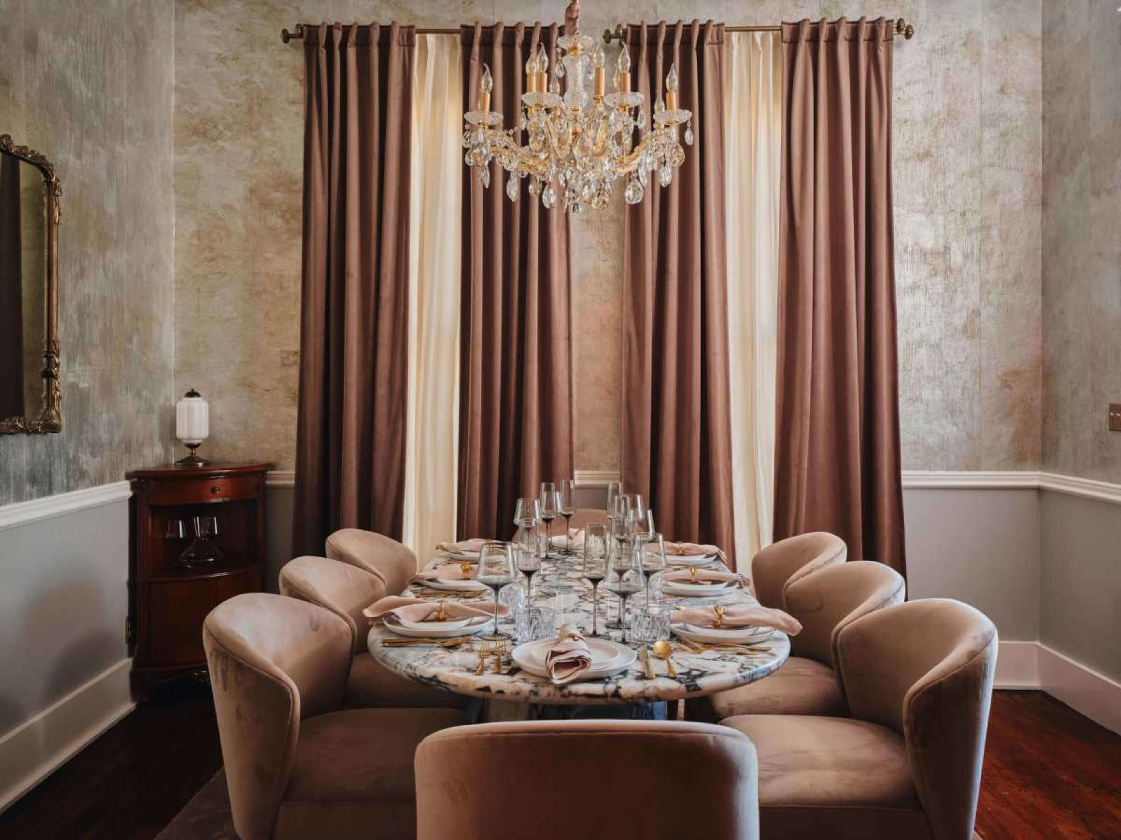 The image shows an elegant dining room with a round marble table set for a meal, featuring plush seating and a chandelier overhead.