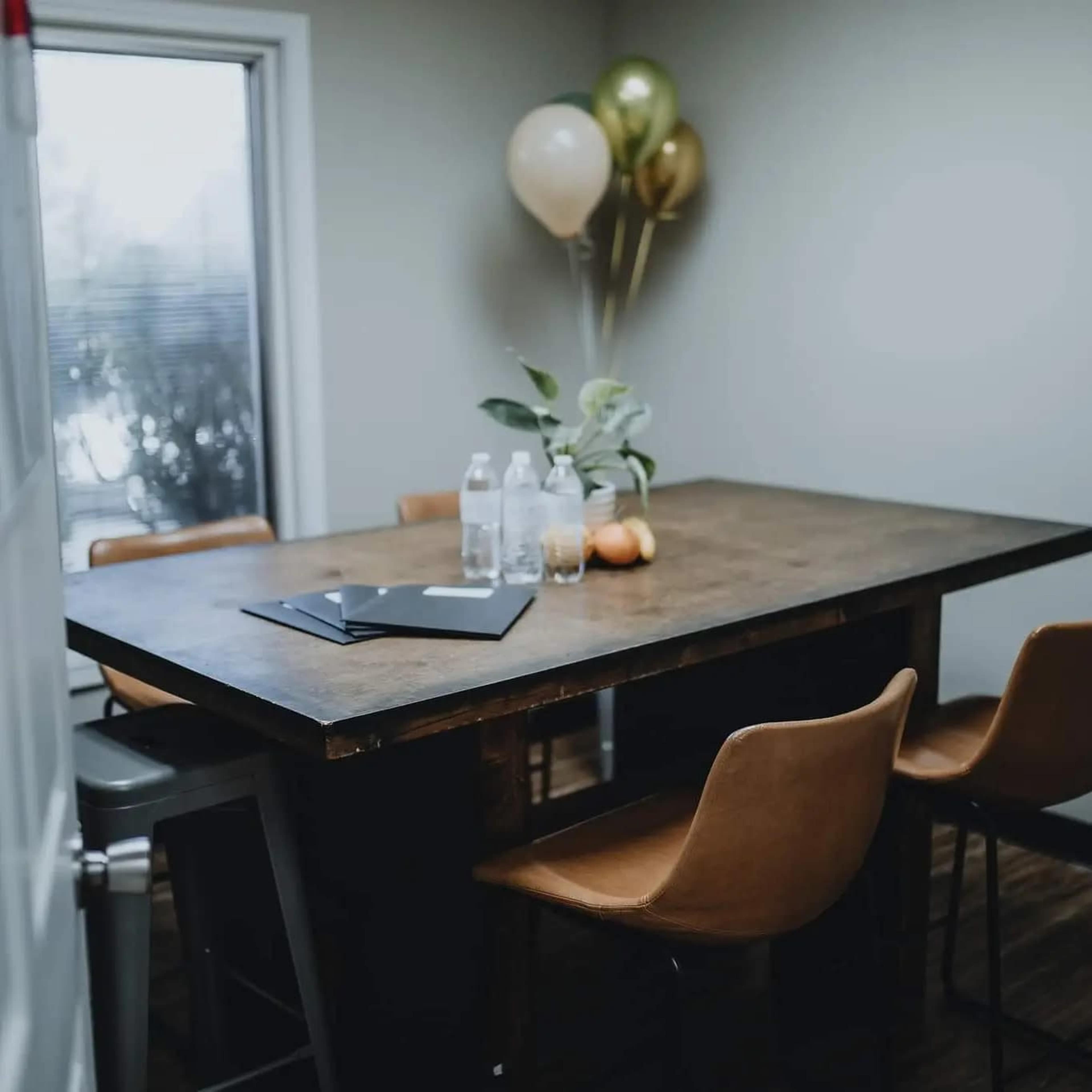 A wooden table is set in a room with a few chairs, decorated with water bottles, a plant, and balloons.