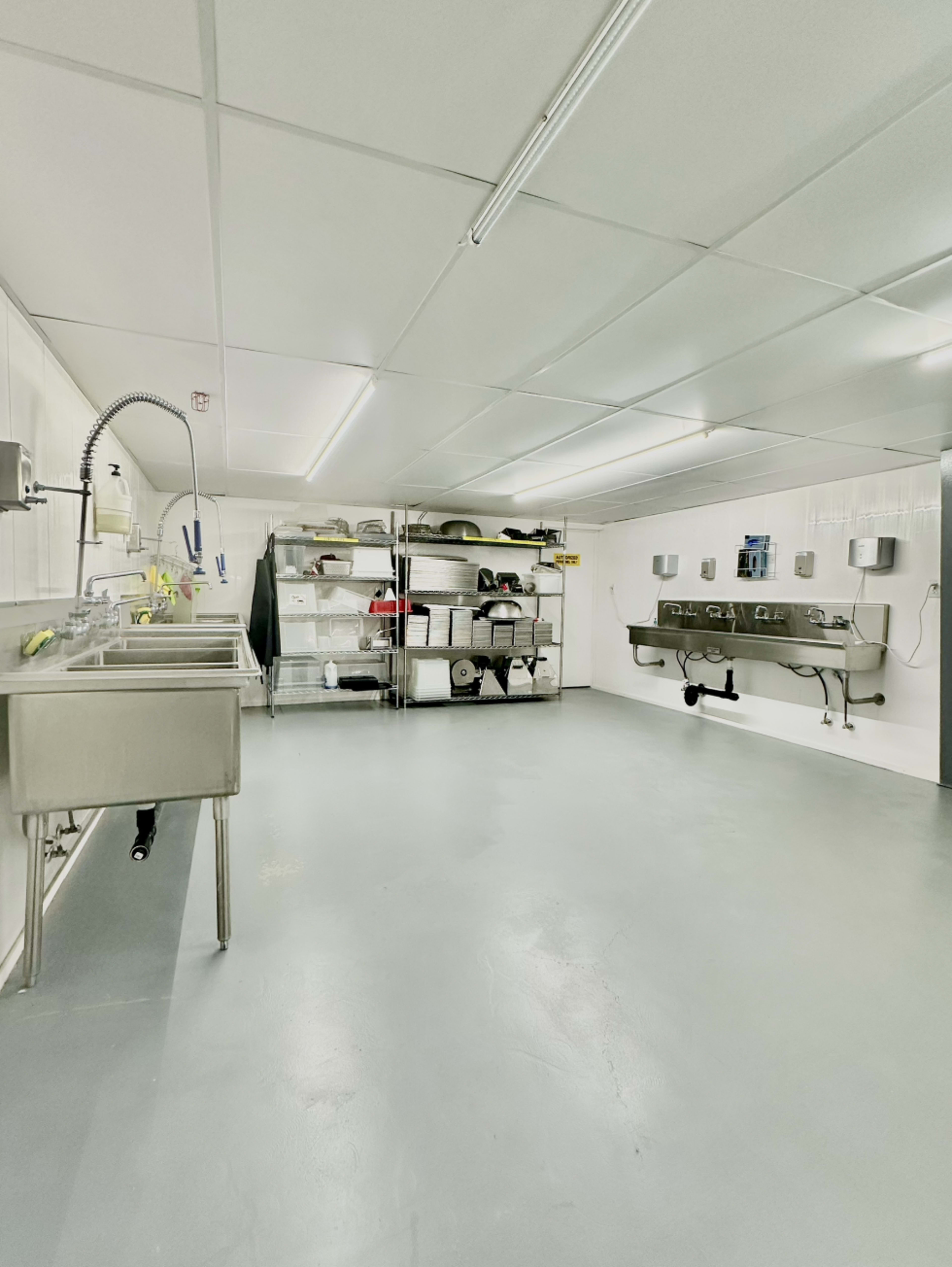 The image shows a clean, spacious kitchen area with stainless steel sinks and metal shelving units against the walls.