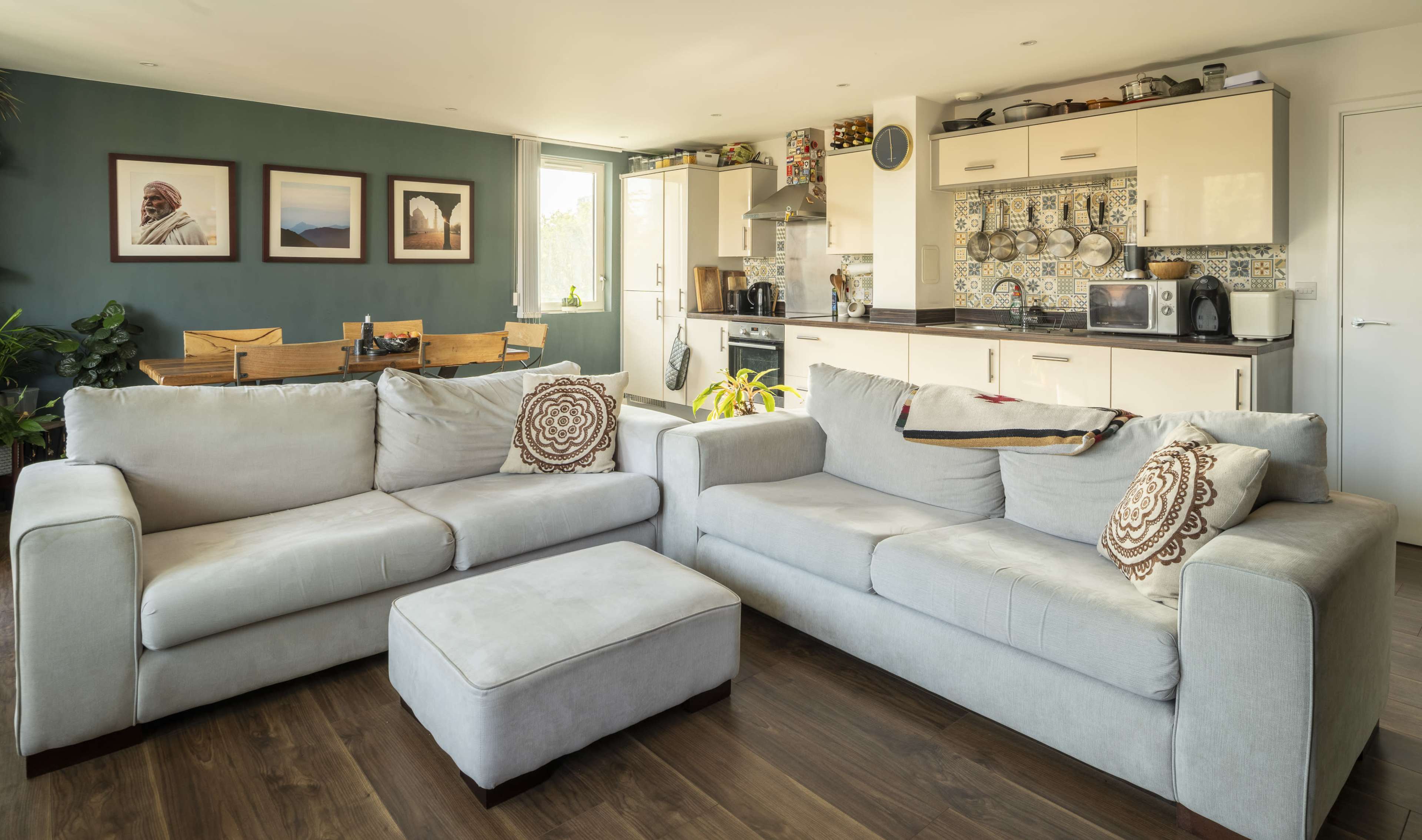 The image shows a modern living room with two light-colored sofas, a coffee table, and an adjoining kitchen area featuring cabinets and a dining table.