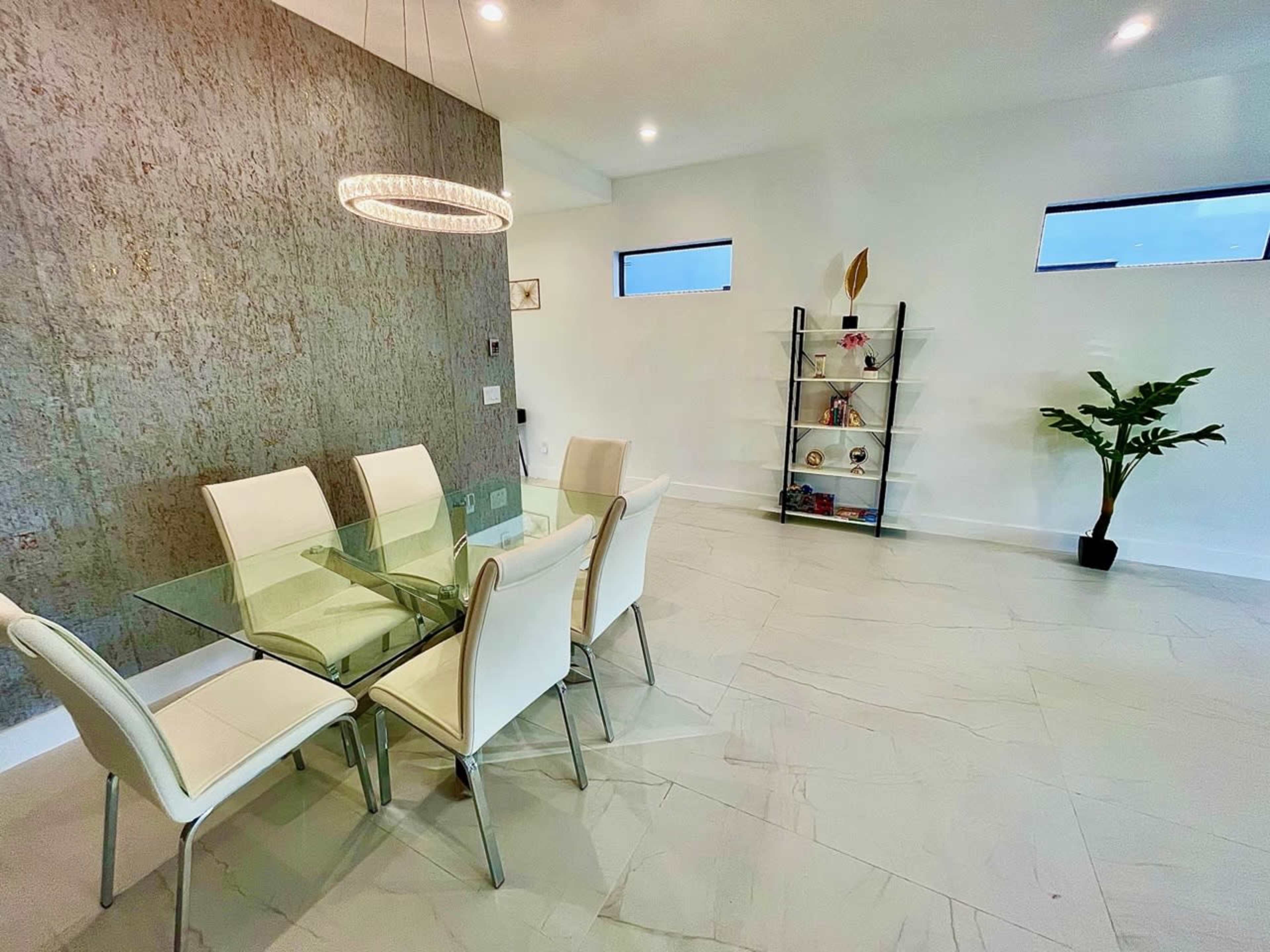 A modern dining area features a glass table surrounded by white chairs, with a decorative shelving unit and a potted plant nearby.