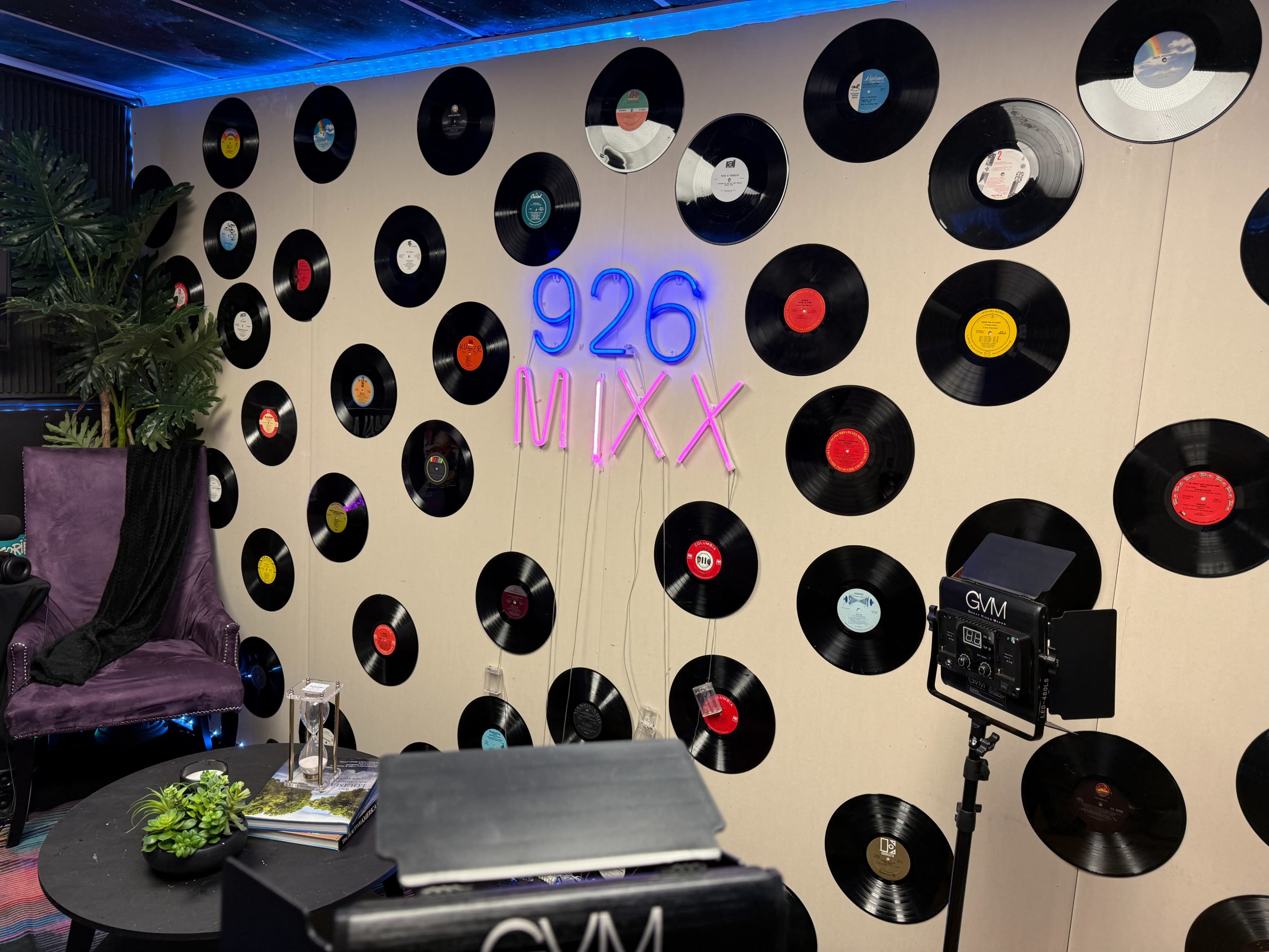 The image features a wall decorated with vinyl records surrounding a neon sign that reads "926 MIXX," complemented by a purple armchair and various items on a black table.