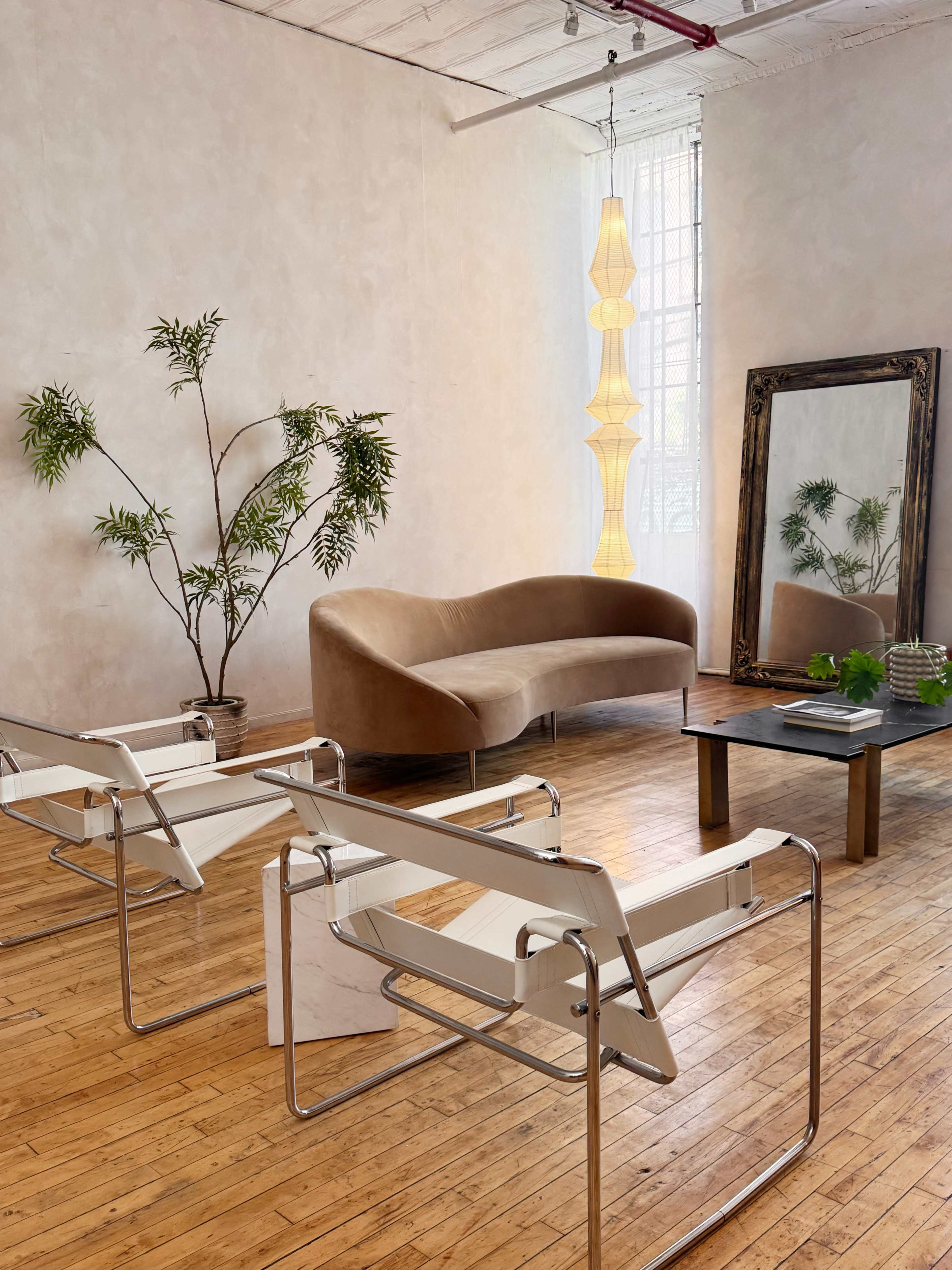 The scene features a well-lit modern living area with a curved sofa, two white chairs, a glass coffee table, a large mirror, and a decorative plant.