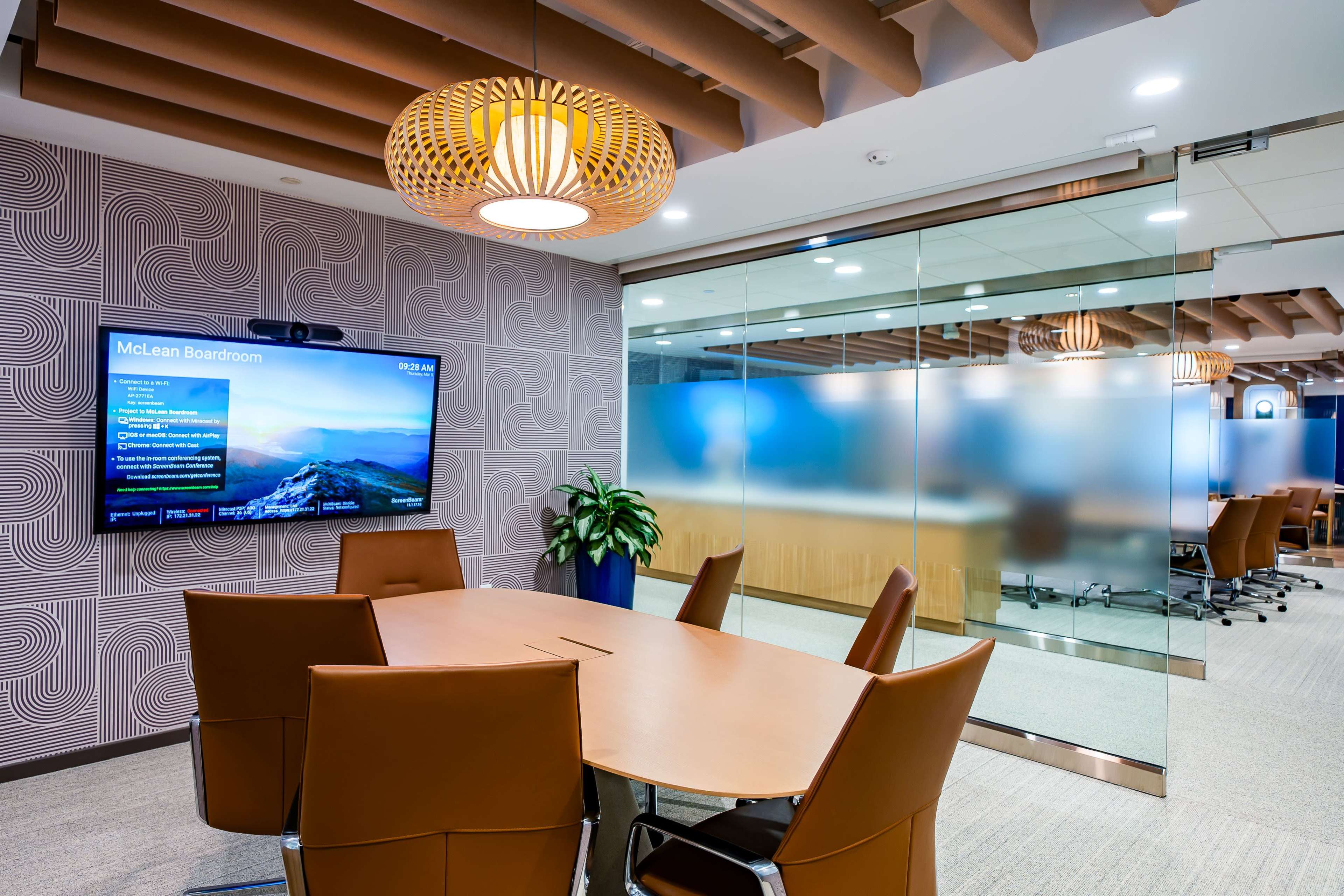 The image shows a modern boardroom featuring a round wooden table, brown leather chairs, a large wall-mounted screen displaying information, and a glass-walled meeting area in the background.