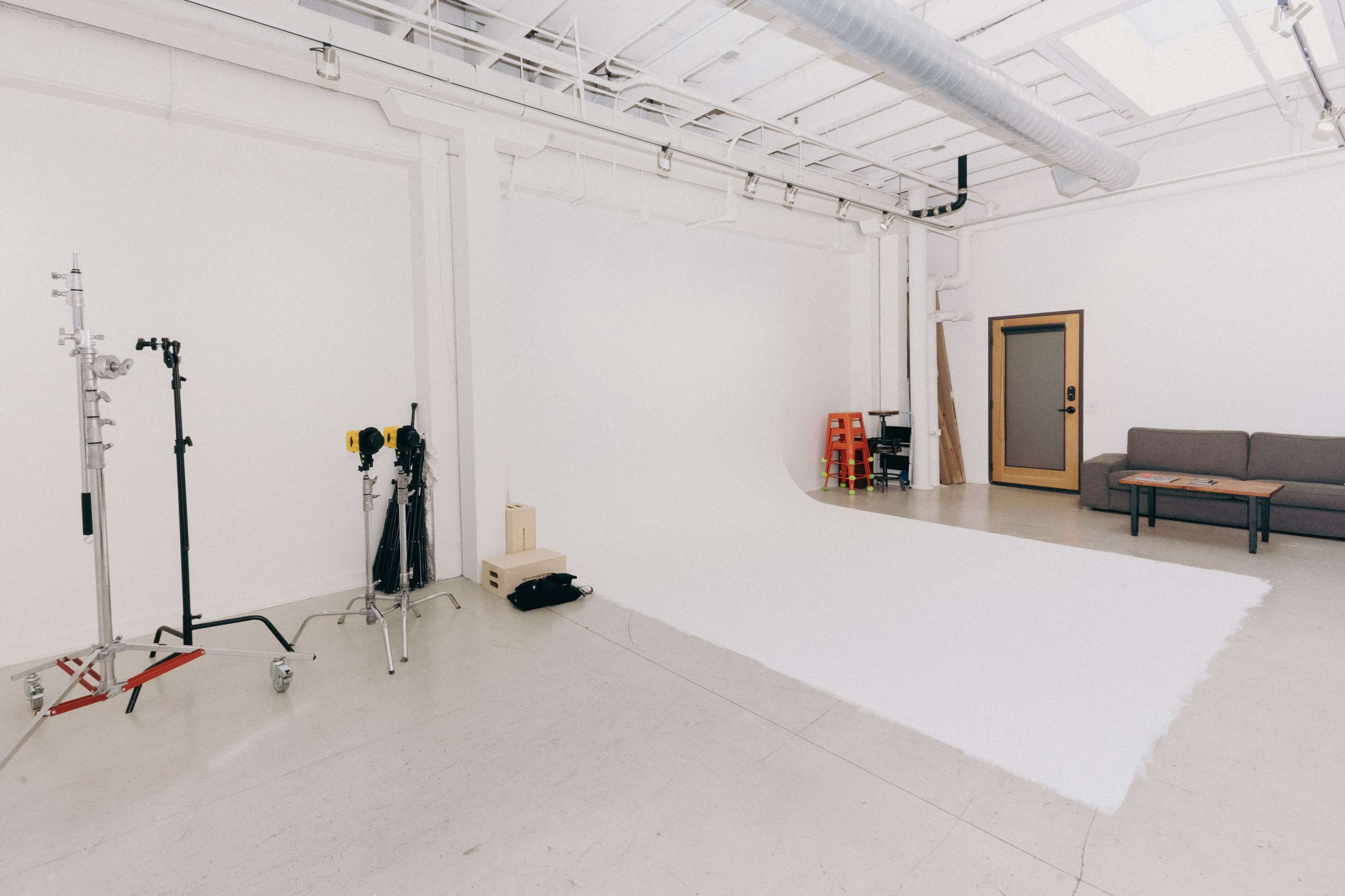 A minimalist photo studio with white walls, a roll of white backdrop material, and a couch next to a door.