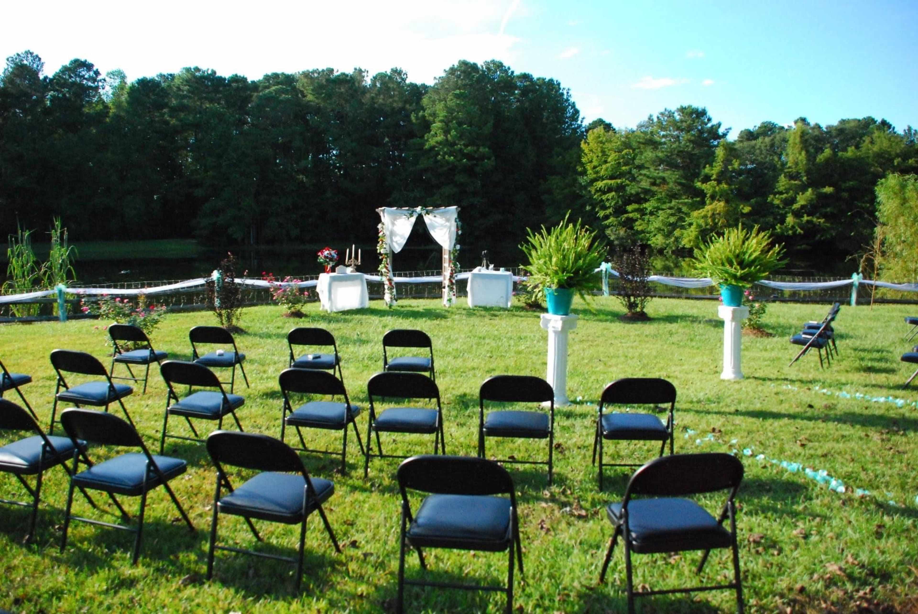 A grassy area set up for a wedding ceremony, featuring rows of black chairs facing a decorated altar with a backdrop of trees and a body of water.