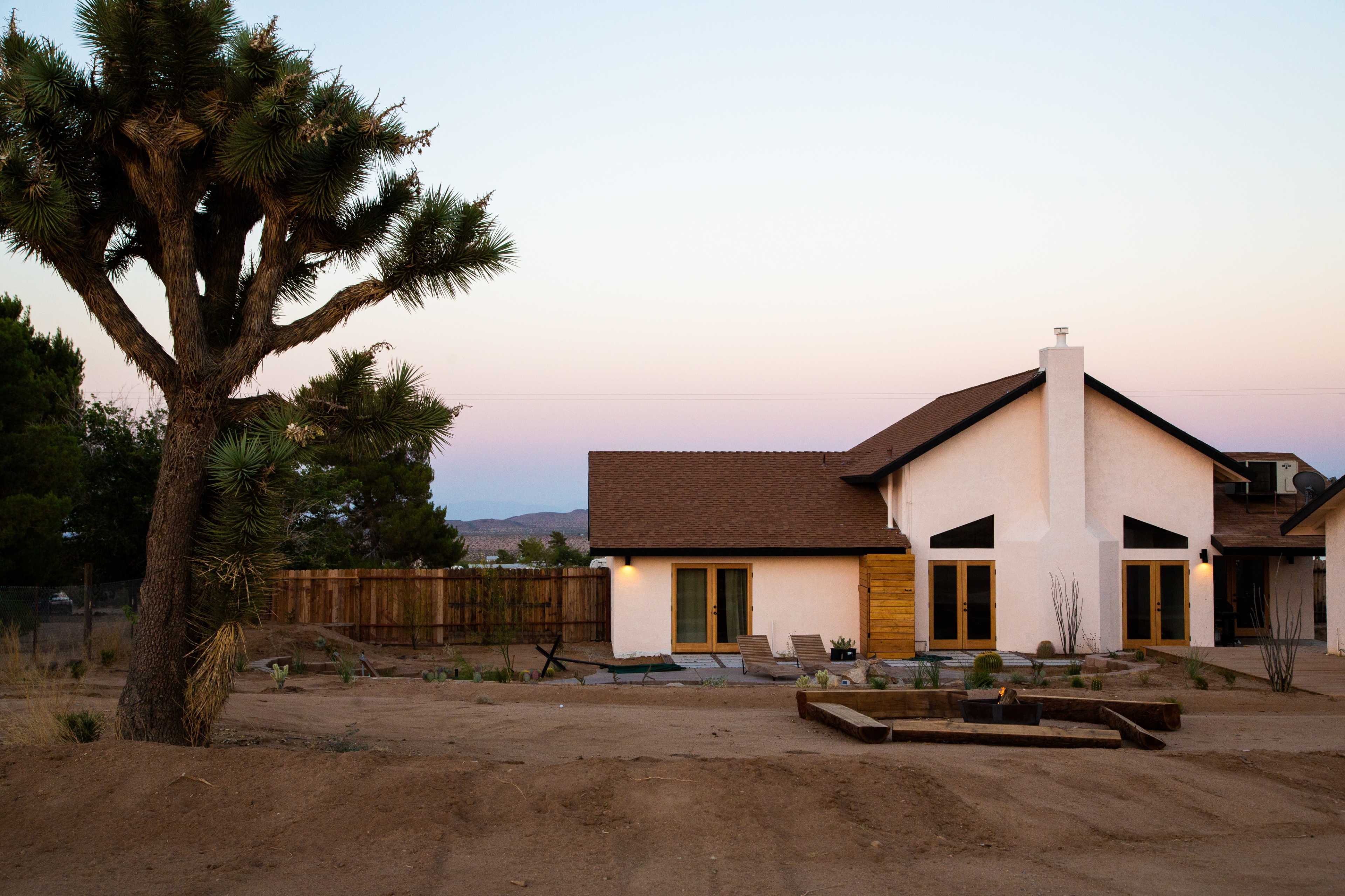 A modern house with a wooden deck and landscaped yard is situated near a large Joshua tree, under a pastel sunset sky.