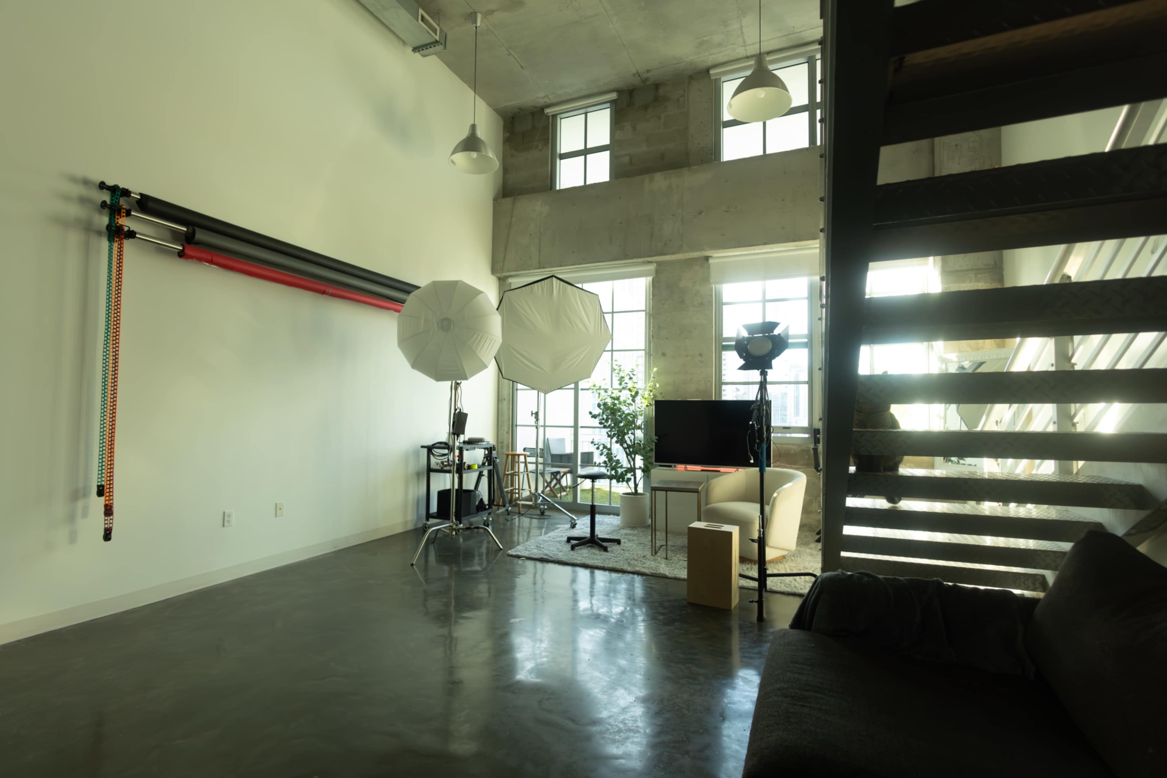 The image shows a spacious, well-lit photography studio featuring softboxes, a staircase, and a seating area with a large window.