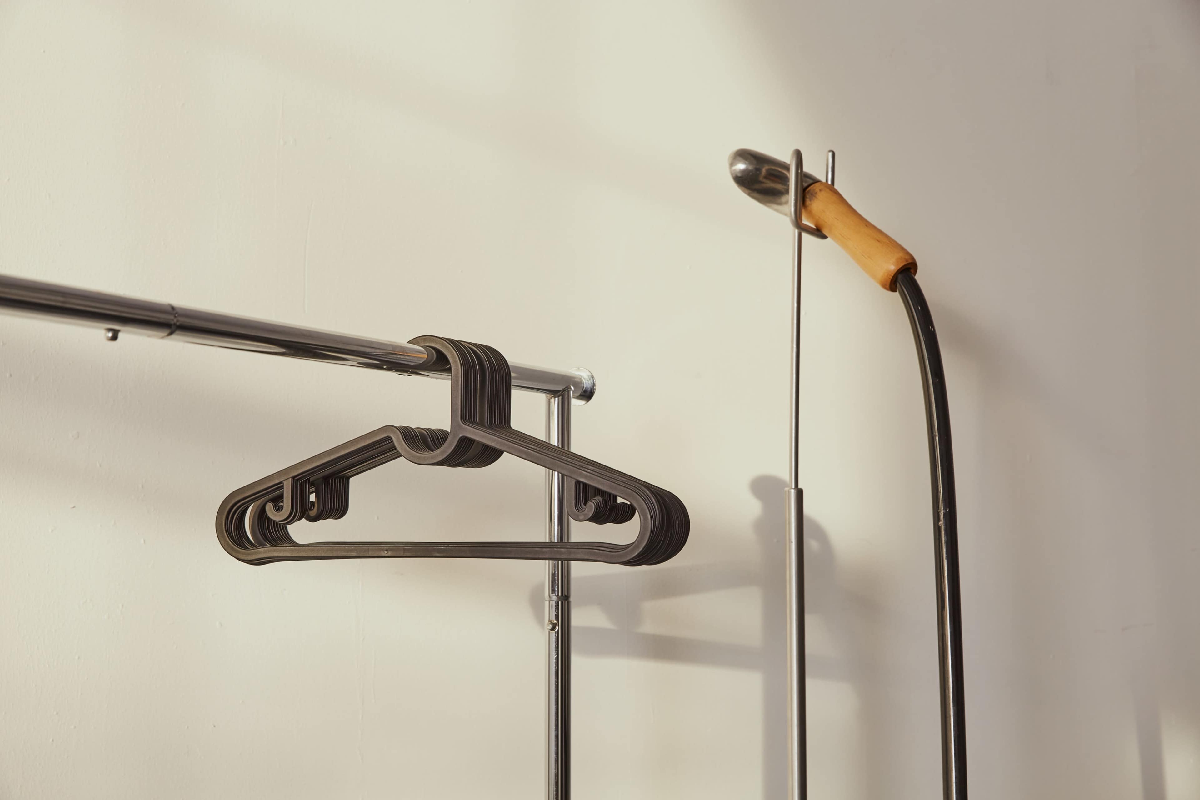 The image shows a metal clothing rack with a black hanger and a garment steamer with a wooden handle against a plain wall.