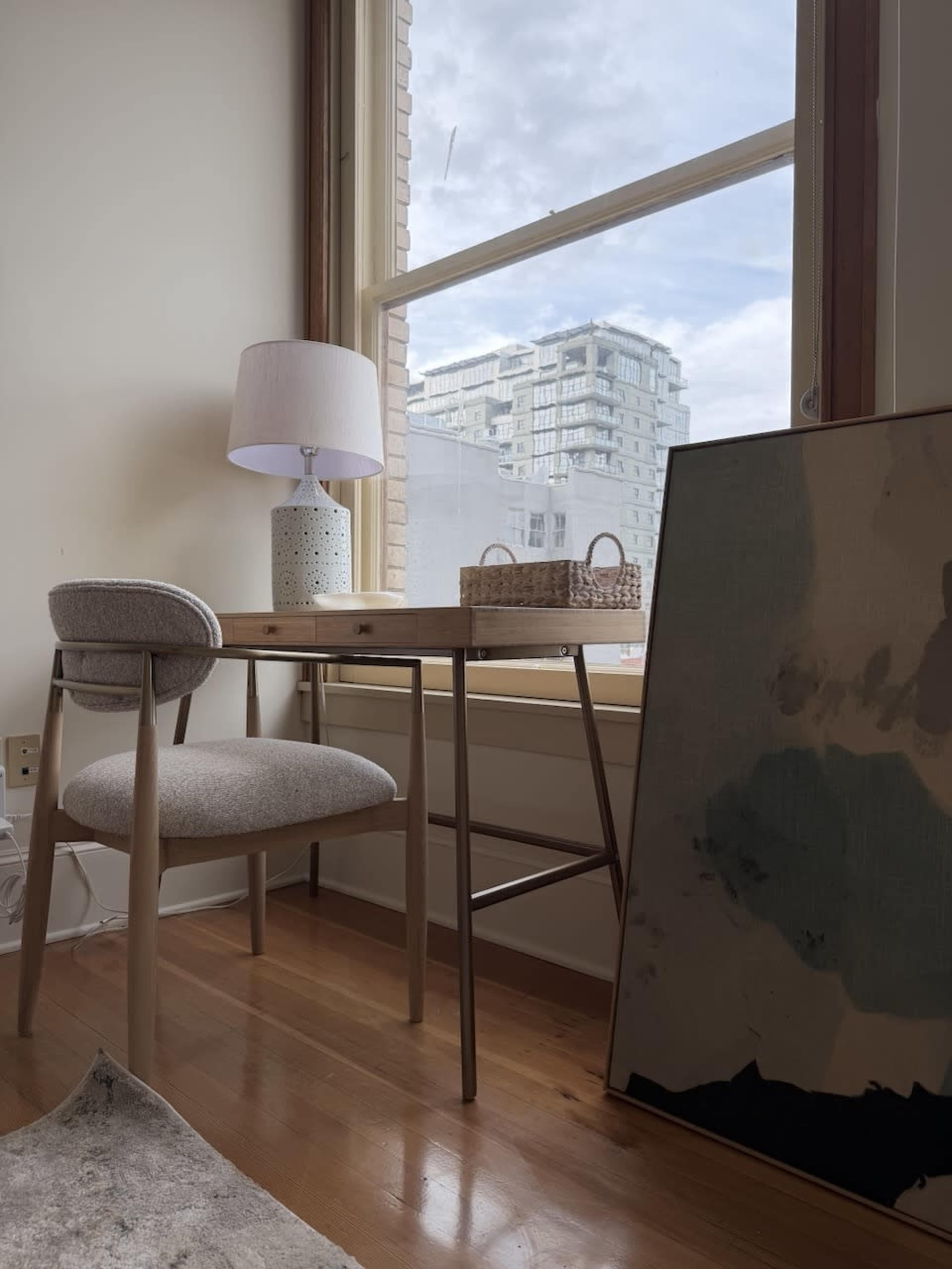 A wooden desk with a lamp and a chair is positioned near a window, overlooking a modern building outside.