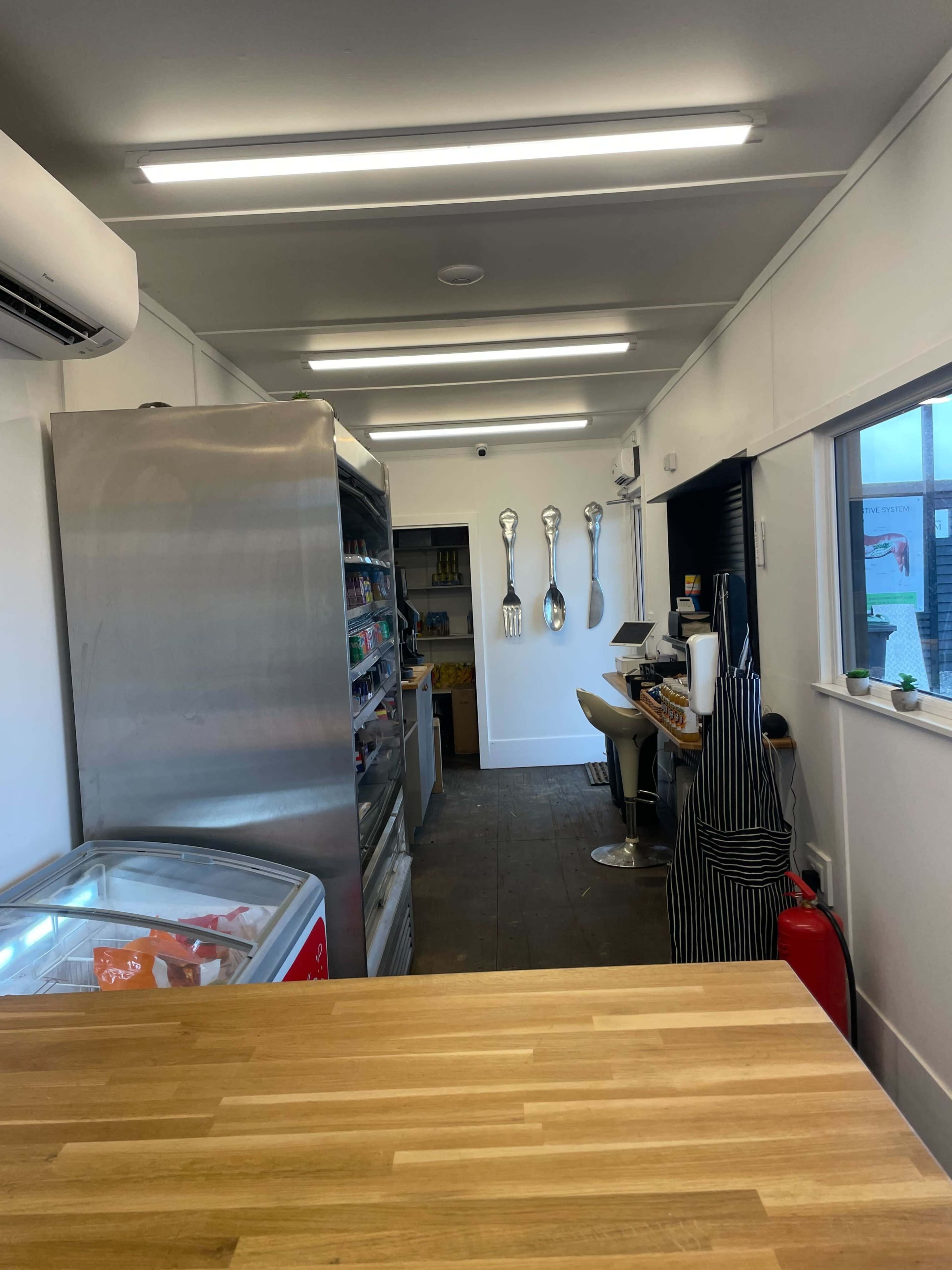 The image shows the interior of a small kitchen or café space featuring a metal refrigerator, shelves stocked with various food items, and large cooking utensils displayed on the wall.