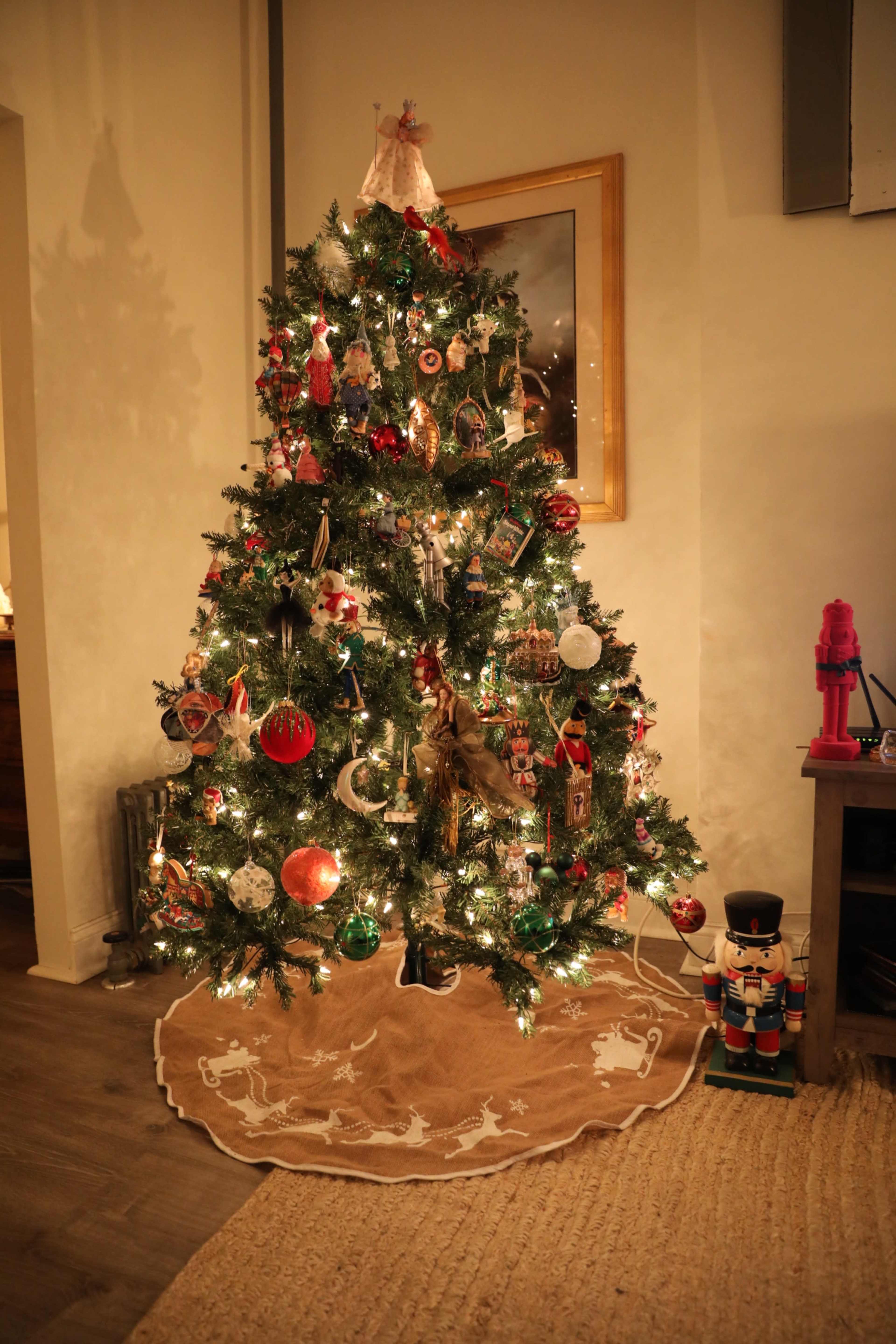 A decorated Christmas tree with colorful ornaments and lights stands in a cozy room, surrounded by a tree skirt and a nutcracker figure nearby.