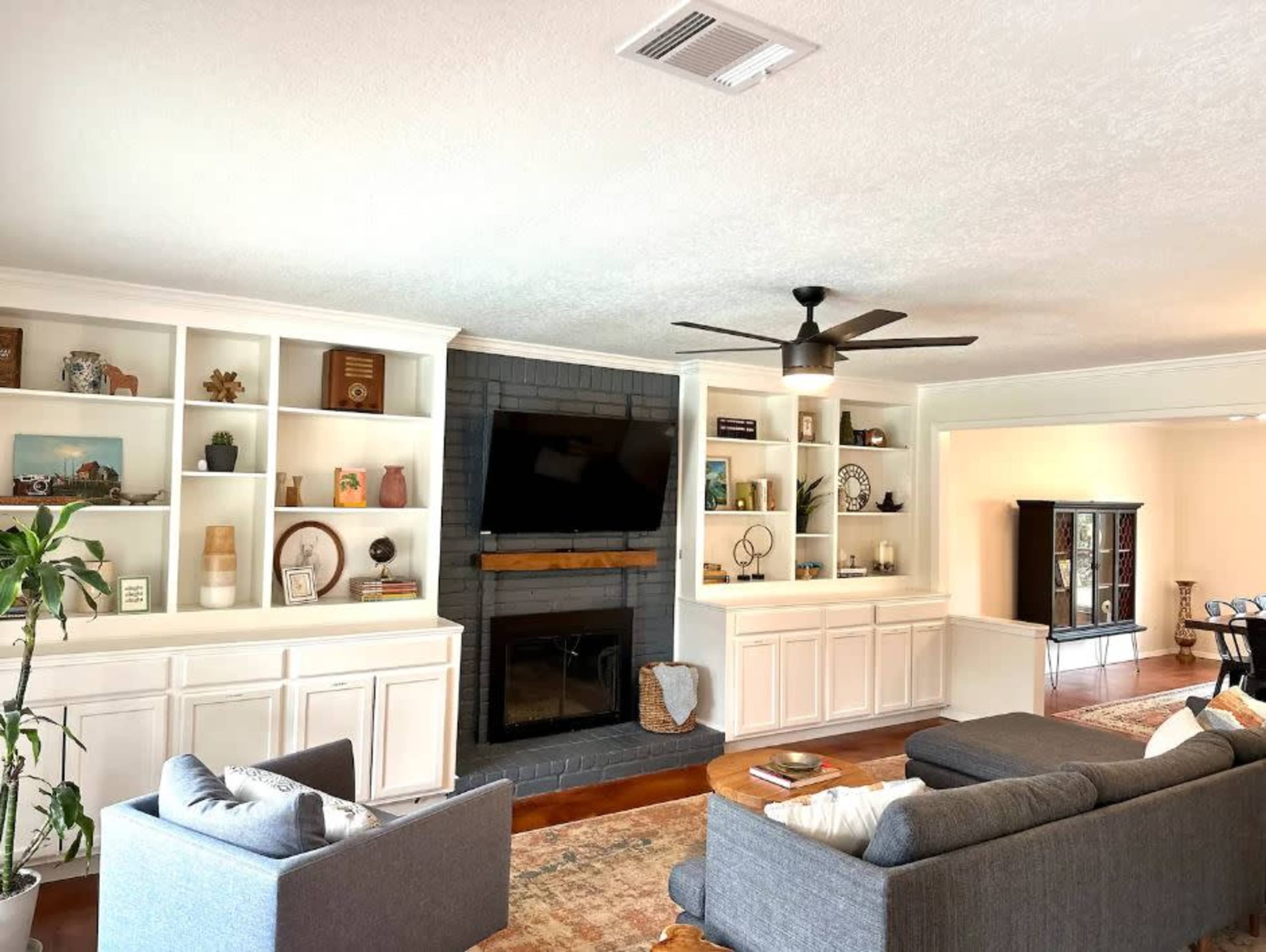 The image shows a living room featuring two gray sofas, a dark brick fireplace, built-in shelving with decorative items, and a ceiling fan.