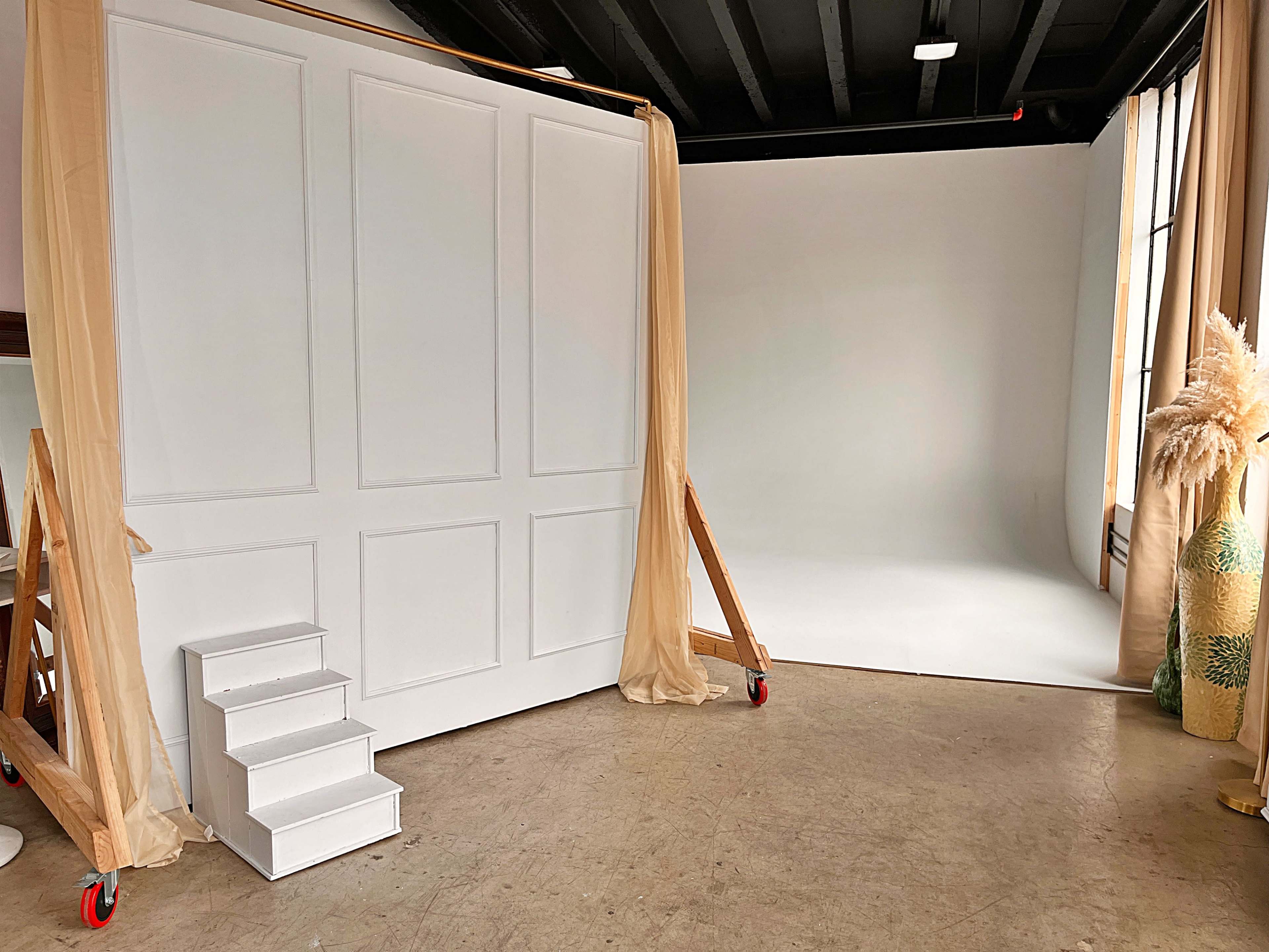 The image shows a photography studio featuring a large white backdrop, a set of steps, and decorative elements, with natural light coming from a window.