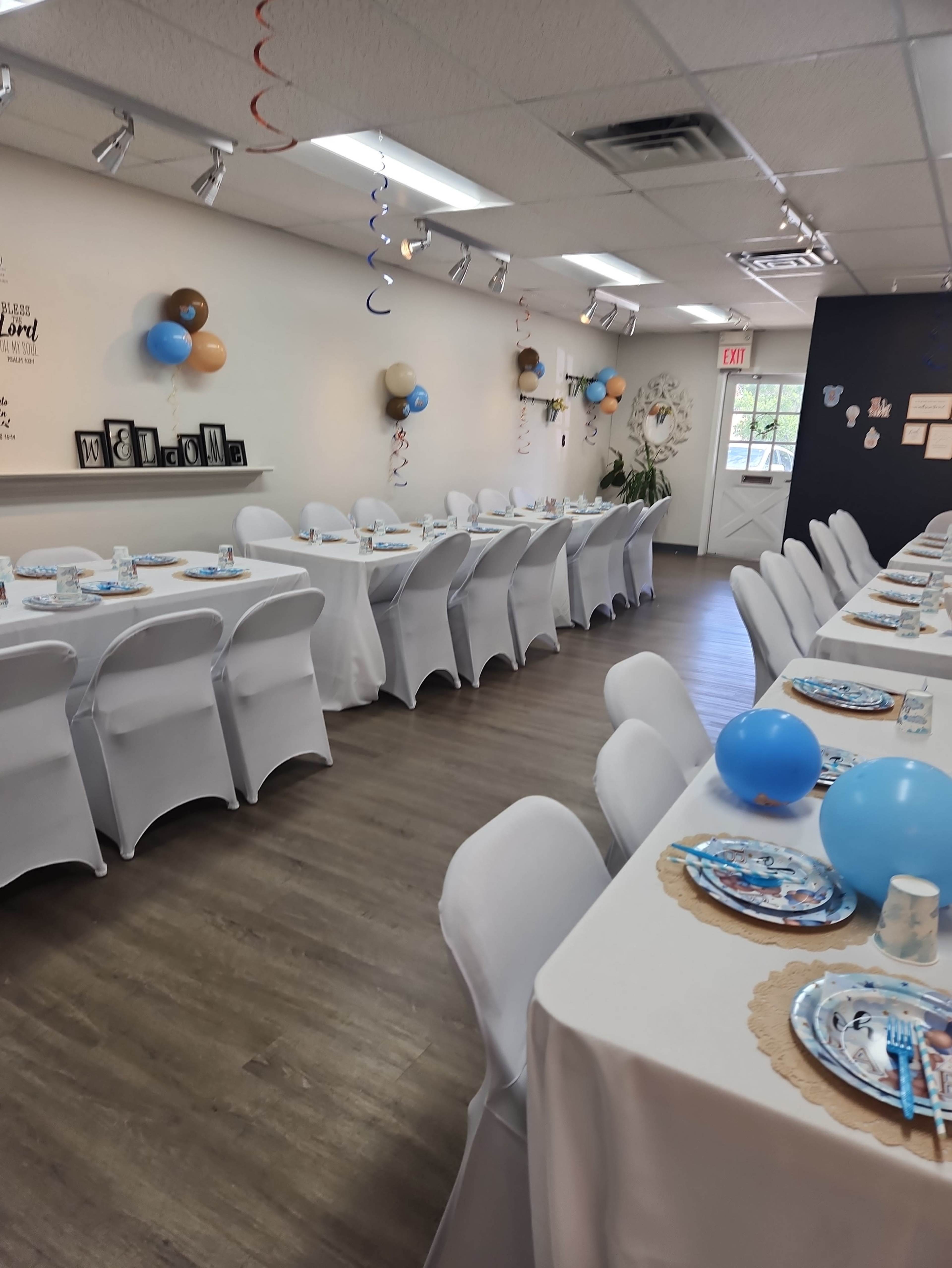 The image shows a decorated party space with two rows of white tables set for an event, featuring blue balloons and decorative place settings.