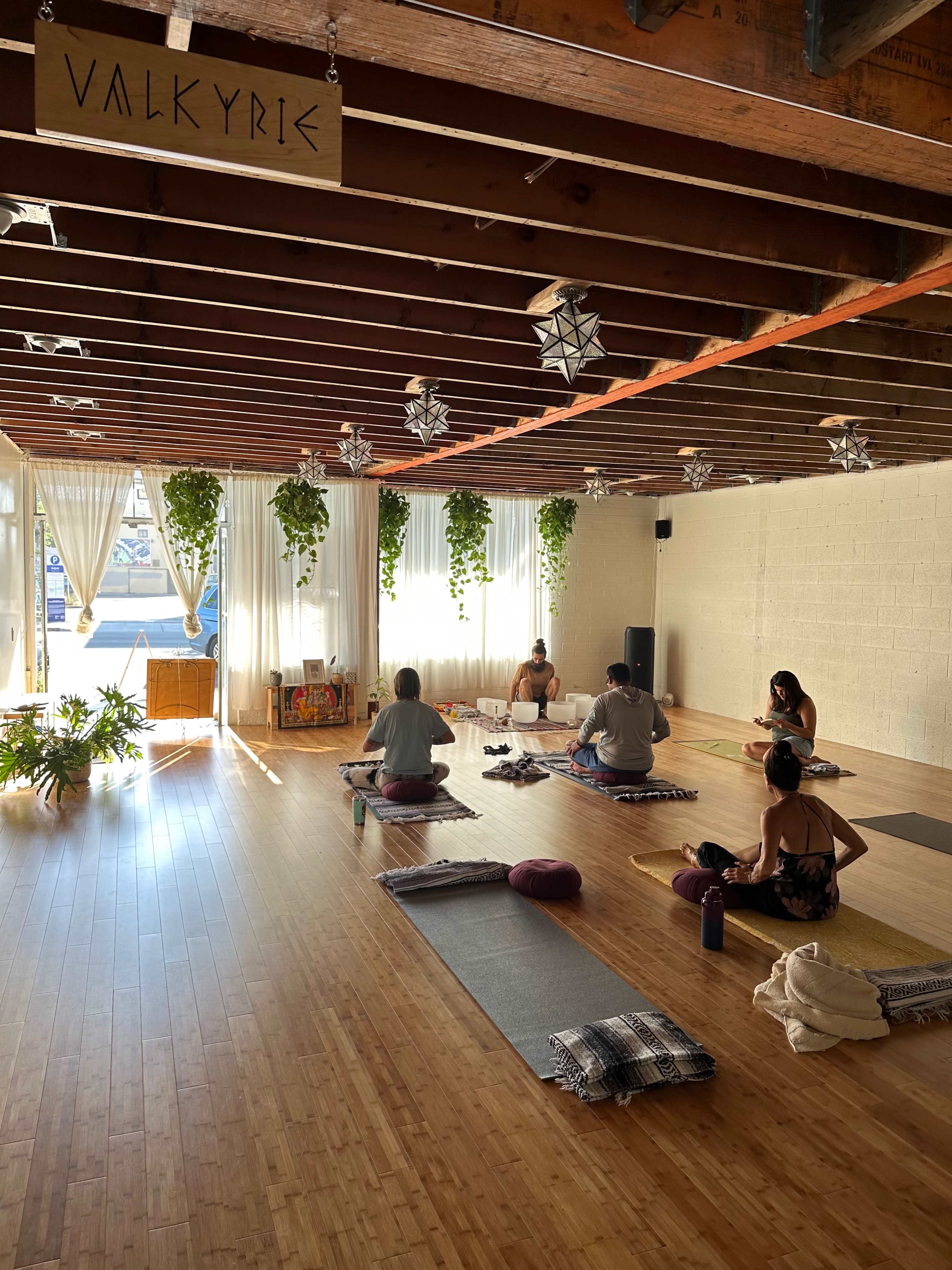 A yoga class is taking place in a spacious studio with wooden floors, natural light filtering through windows, and plants hanging from the ceiling.