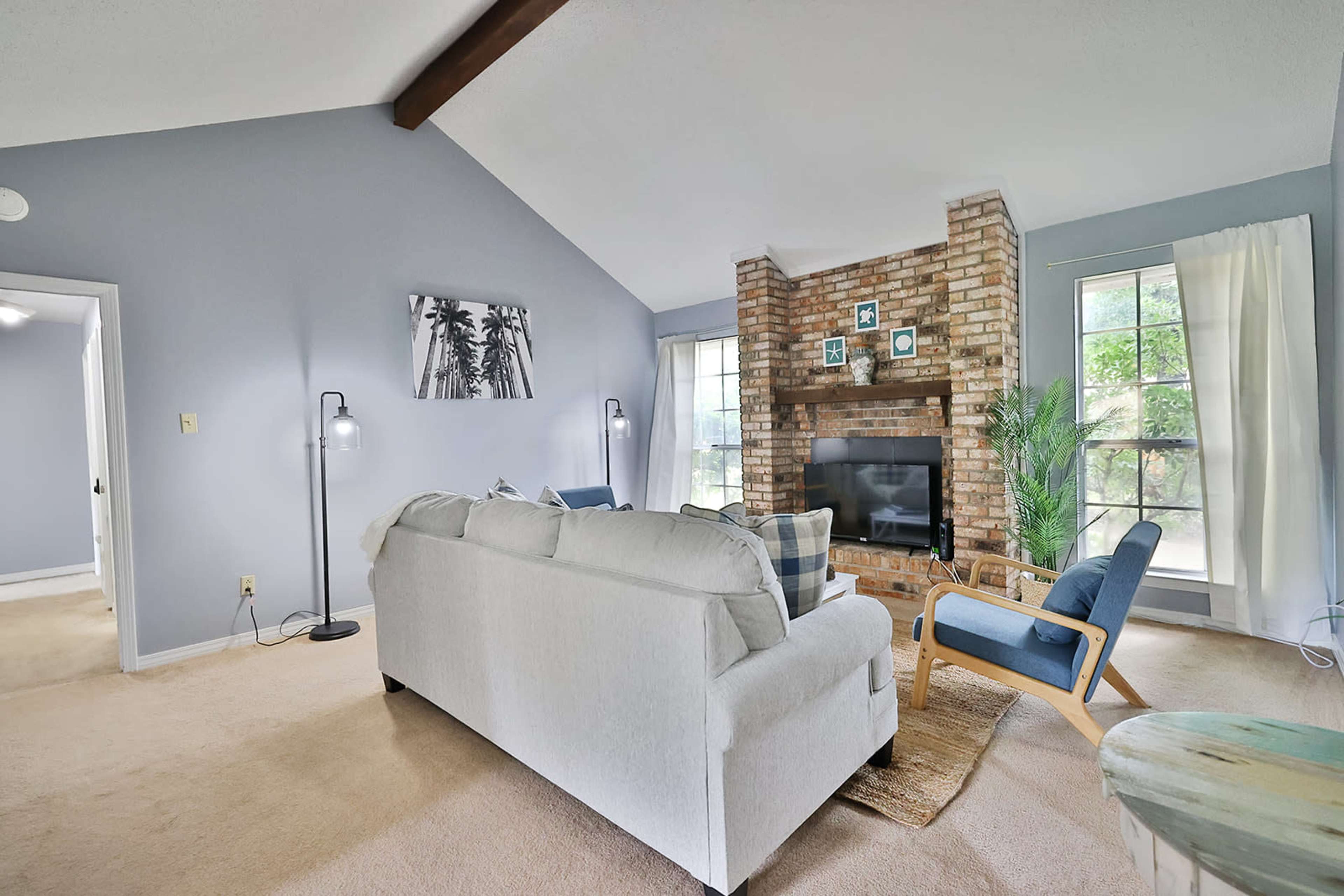 A living room featuring a white sofa, blue accent chair, and a brick fireplace, with large windows and light-colored walls.