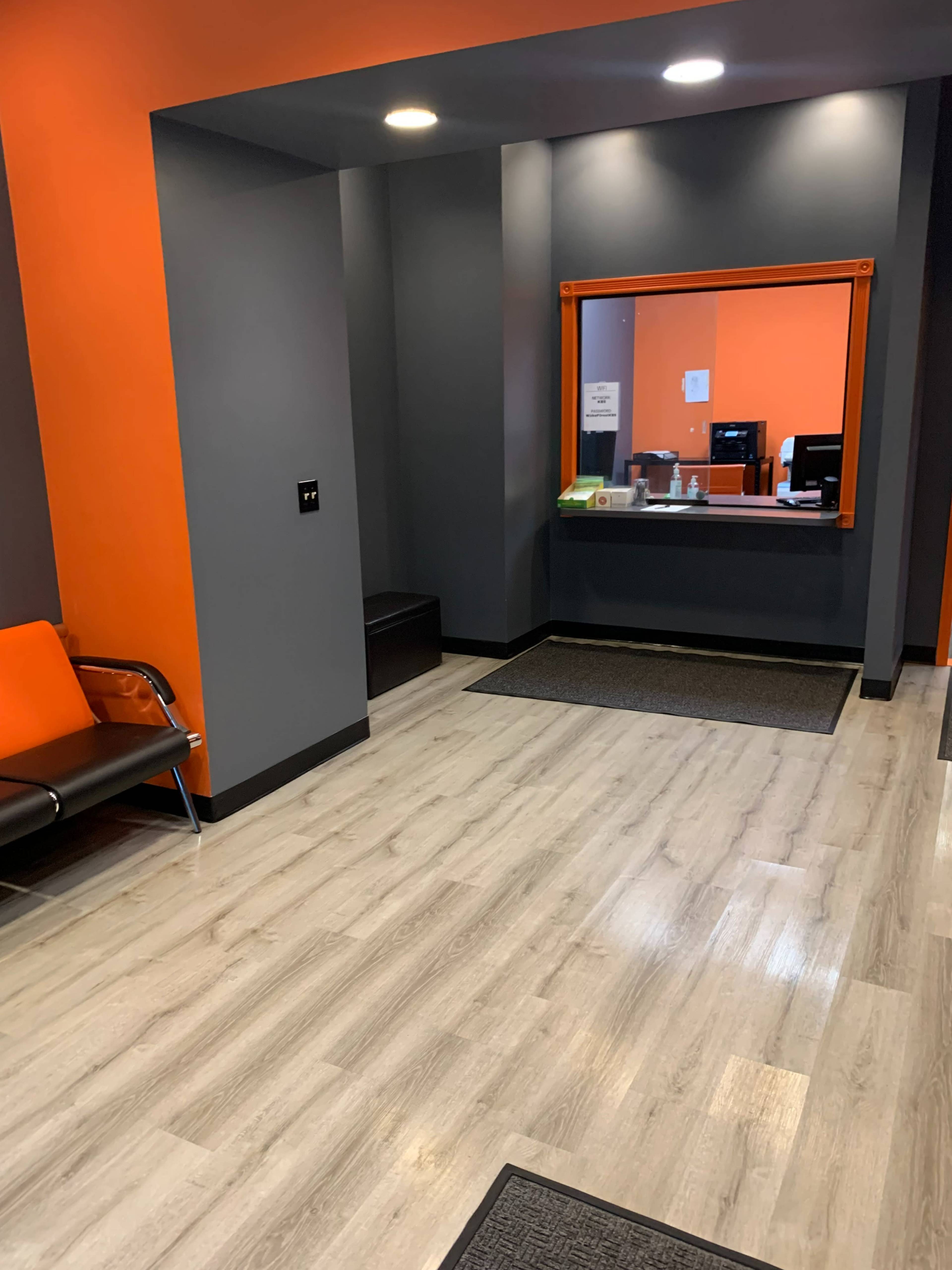 A modern reception area with gray walls, orange accents, a seating area, and a window counter leading to a workspace.