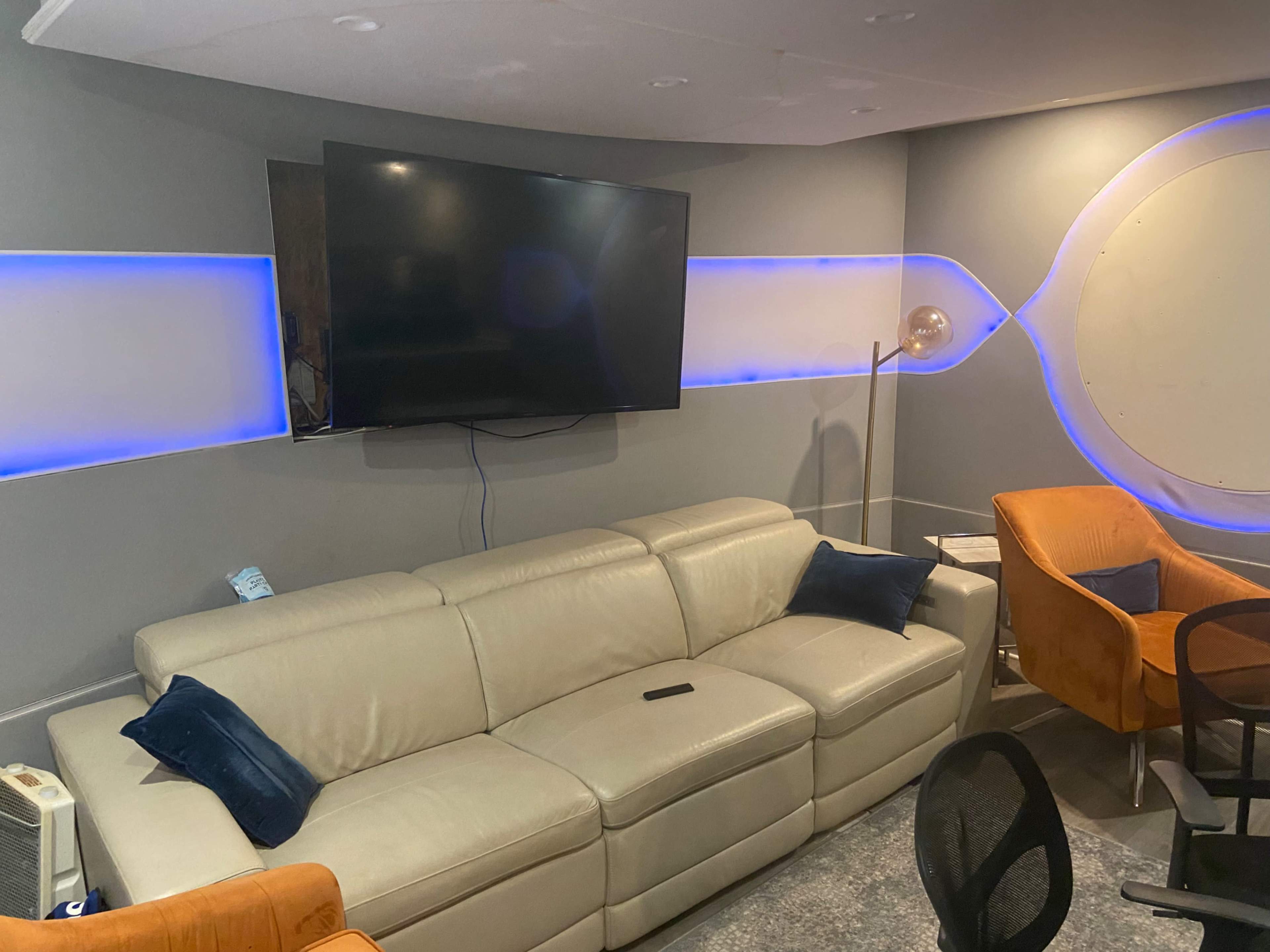 A modern, minimalist living room with a sectional beige sofa, a large flat-screen TV mounted on the wall, and two orange armchairs.