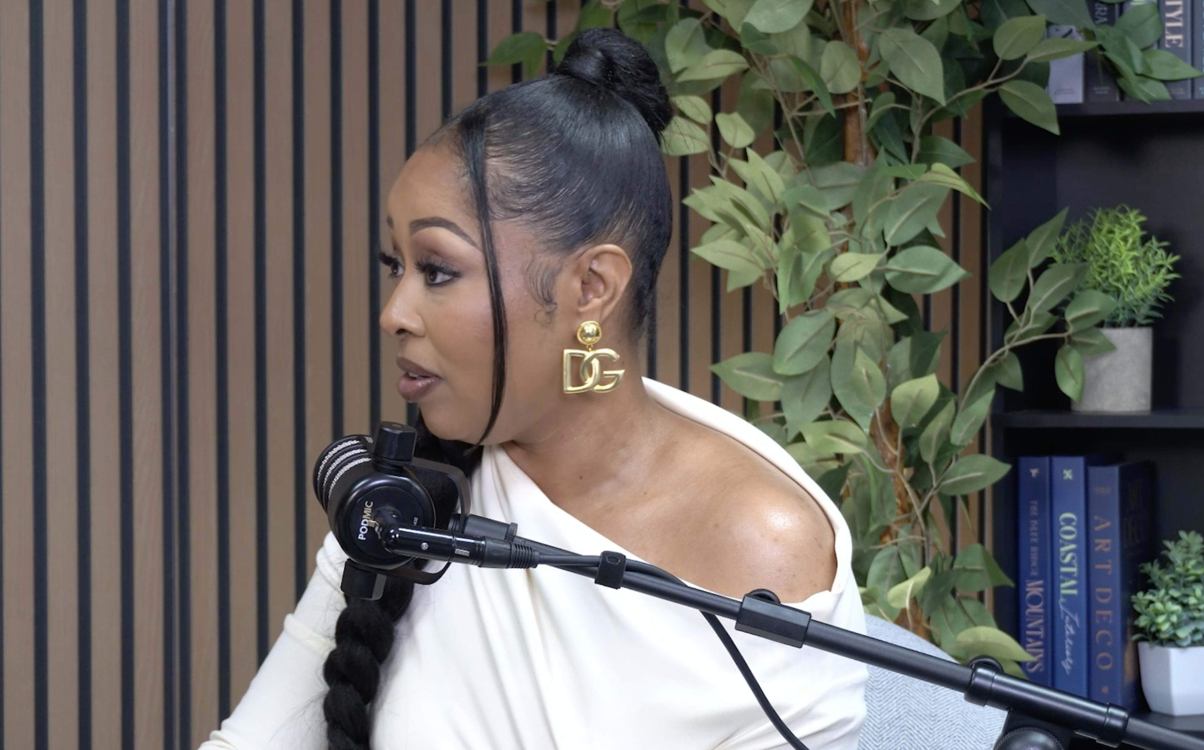 A woman with a ponytail hairstyle wearing large earrings is speaking into a microphone in a studio setting with green plants and shelves in the background.