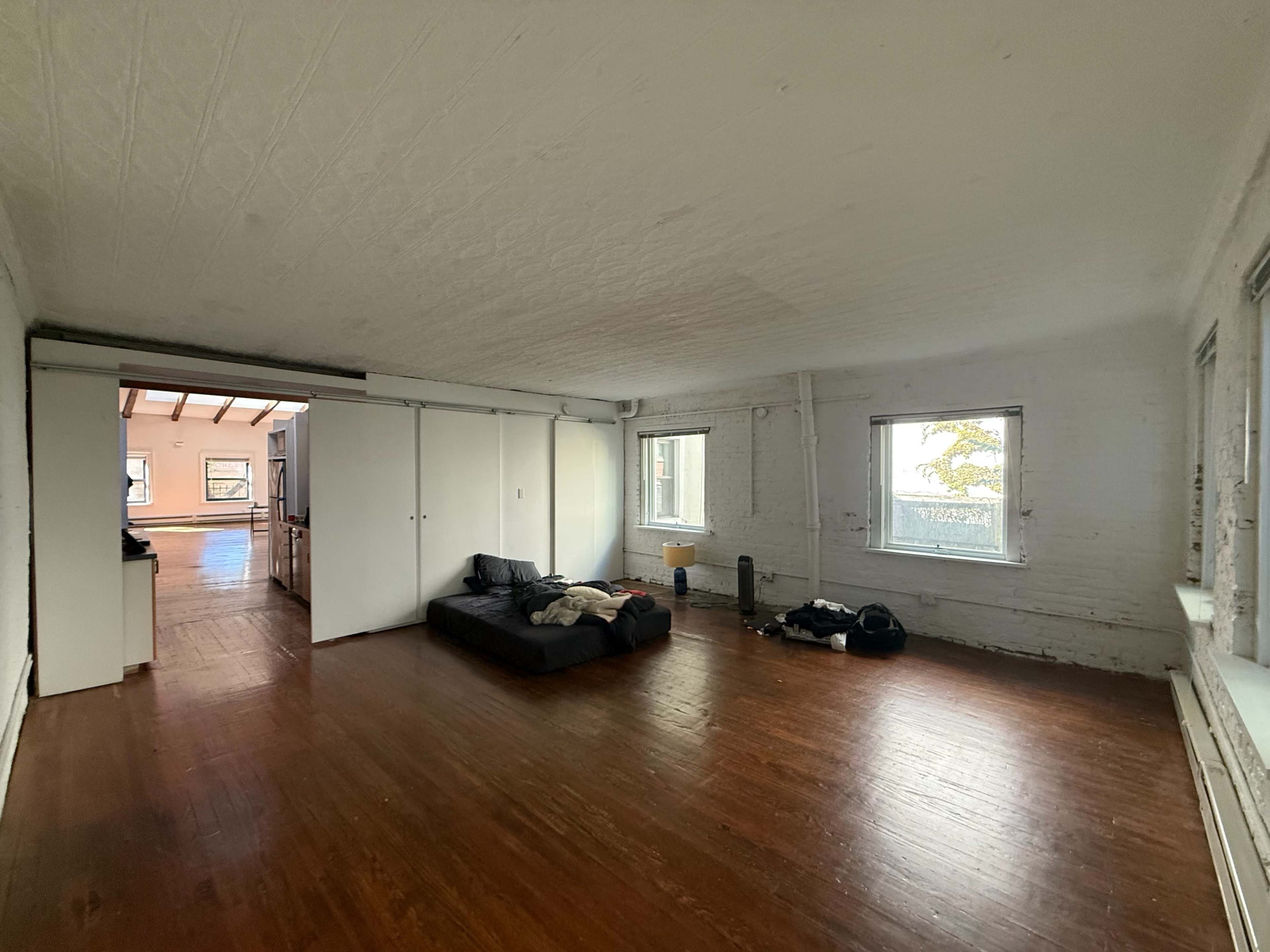 The image shows a spacious, empty room with hardwood floors, white walls, and a black sofa, featuring large windows that provide natural light.