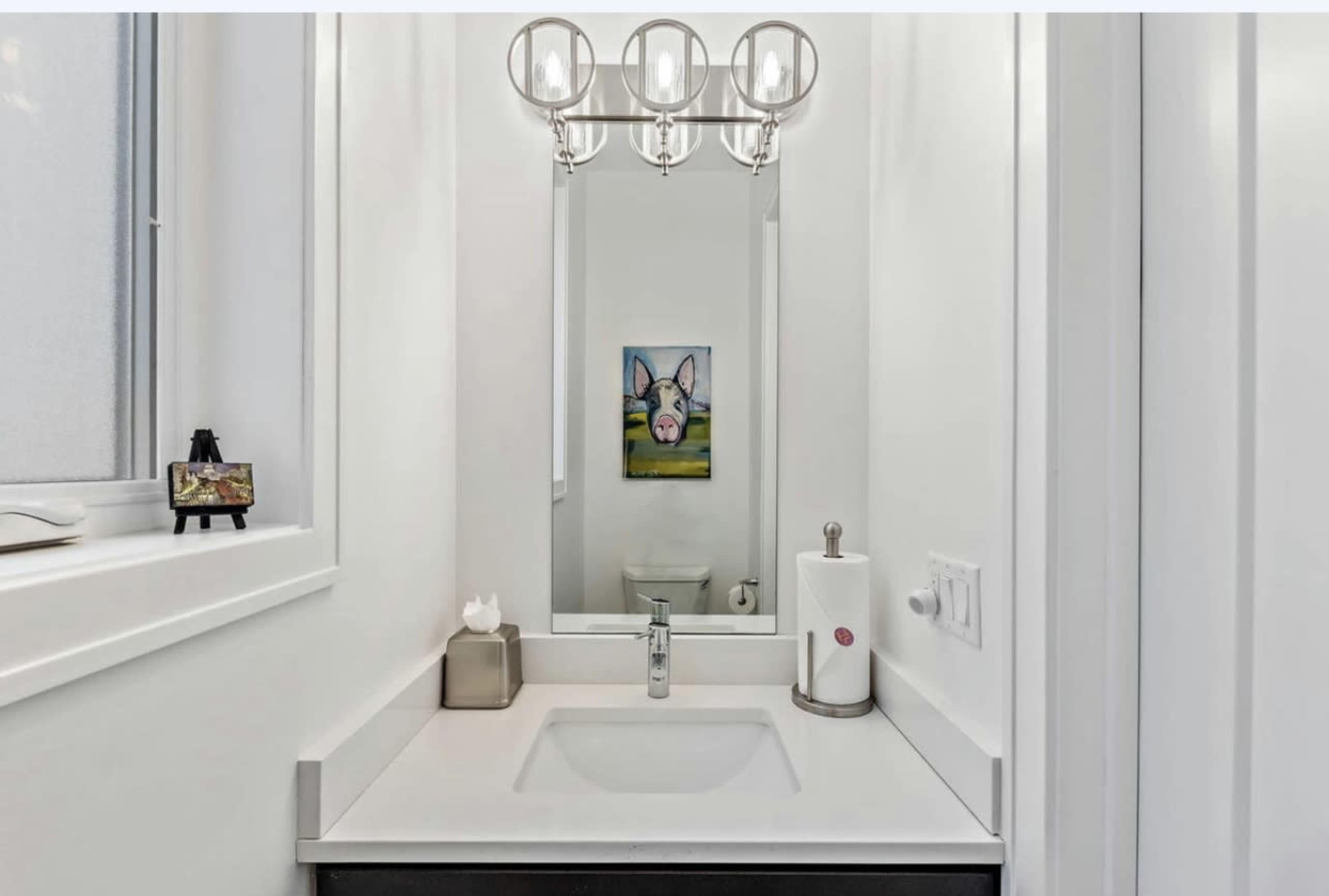 A modern bathroom with a sink, mirror, light fixture, and a painting on the wall.