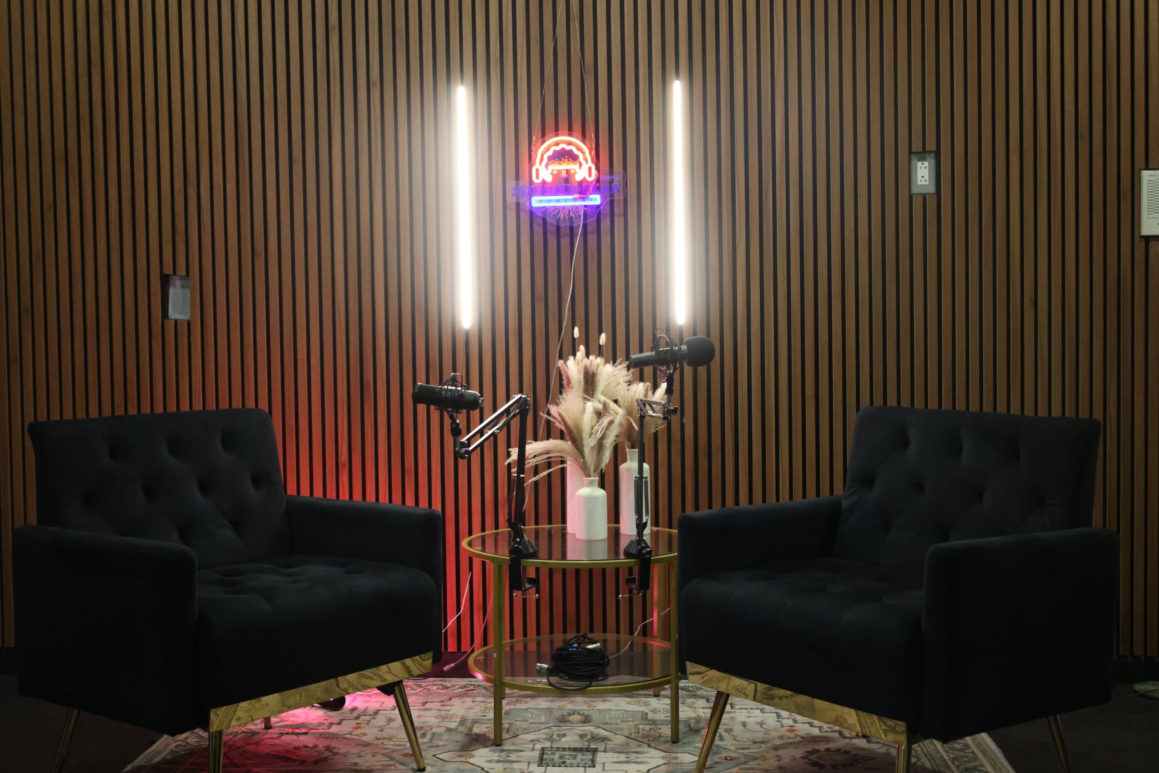 The image shows a podcast studio setup with two black velvet chairs facing each other, a small round table with a plant, and neon lights in the background.