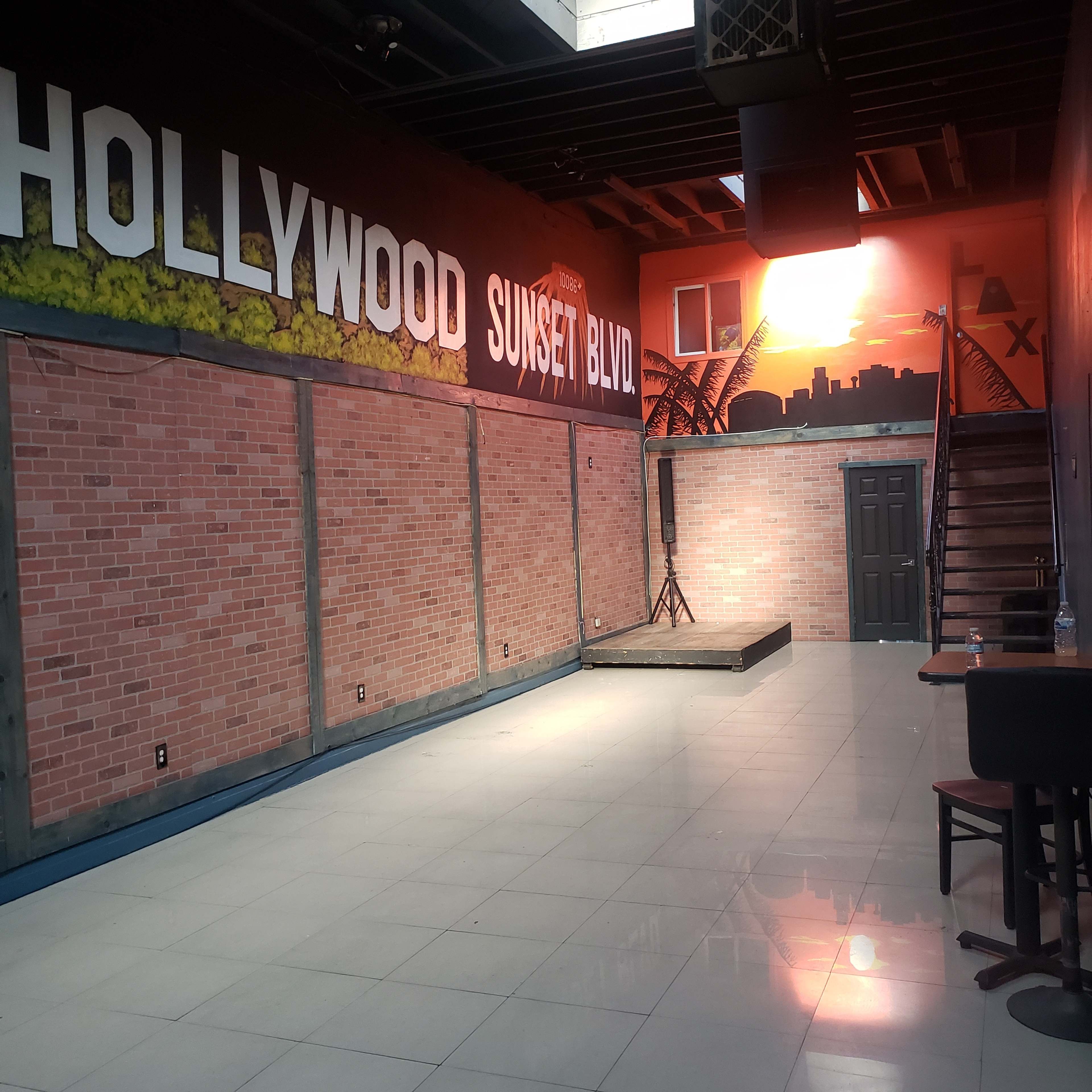 The image shows an empty performance space with a stage, muraled walls featuring "HOLLYWOOD" and "SUNSET BLVD," and a staircase leading to an upper level.