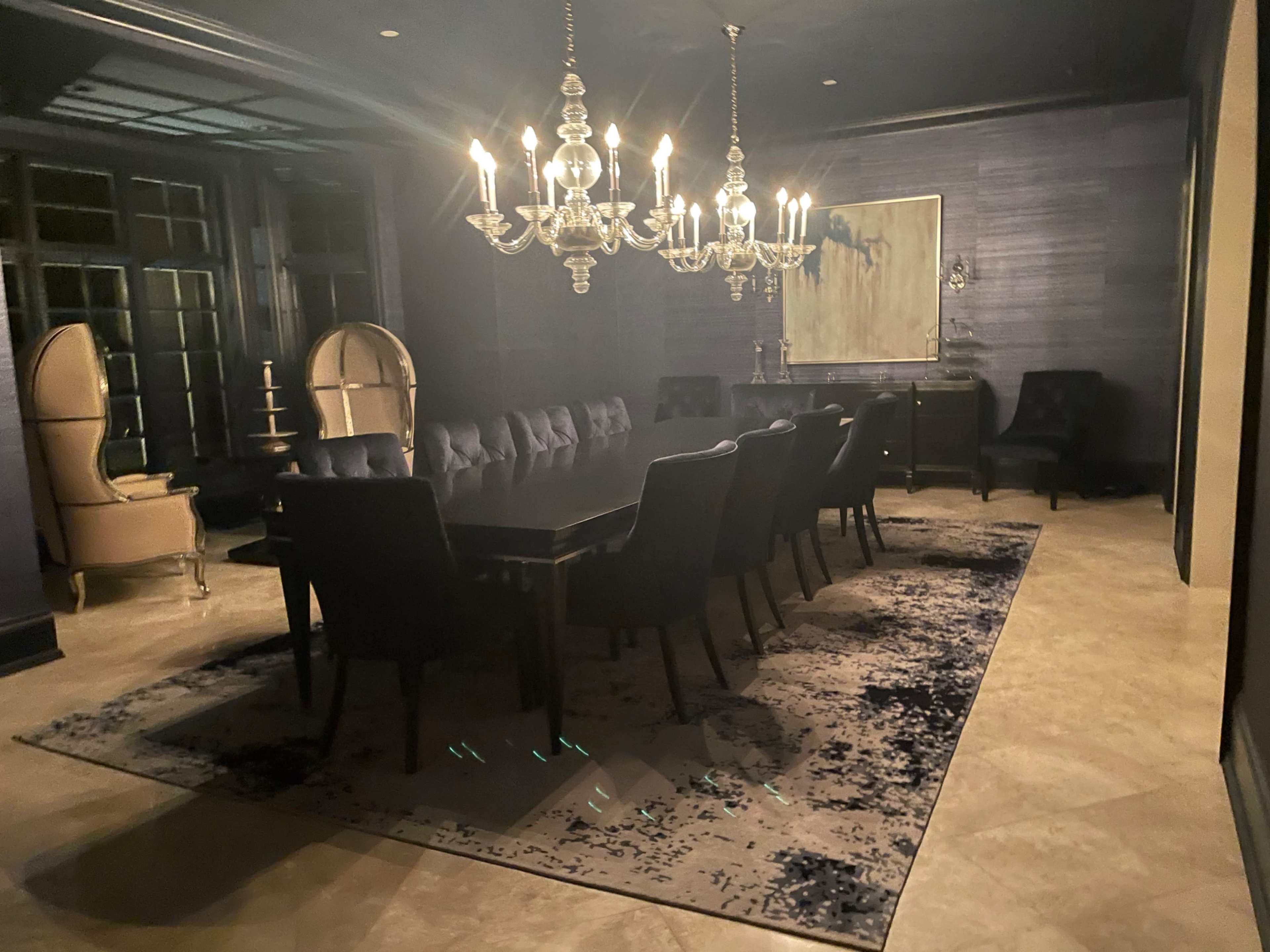 A dark dining room features a long table surrounded by chairs, illuminated by a chandelier and a large rug beneath.
