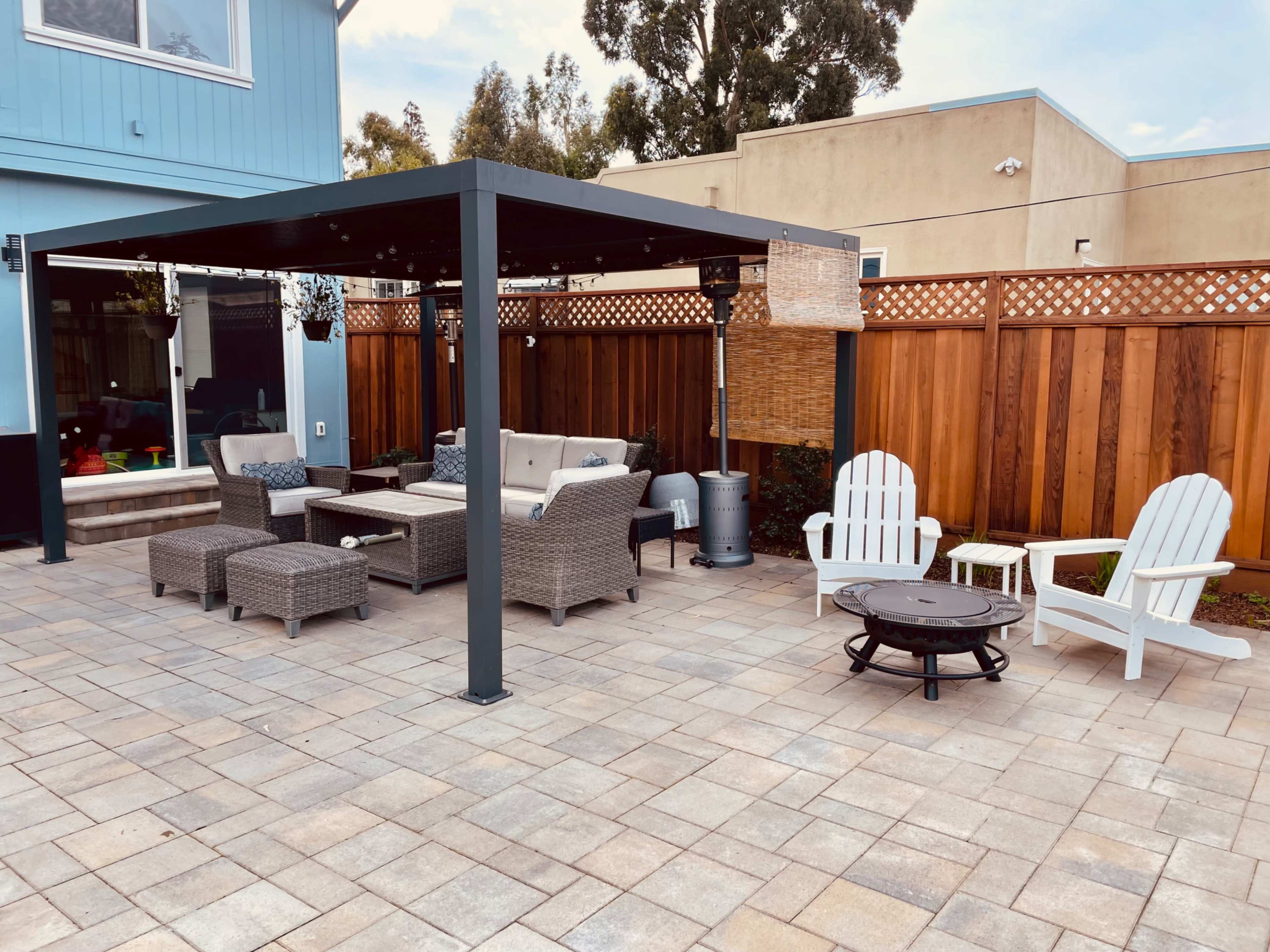 A shaded outdoor seating area features a sectional sofa, chairs, and a fire pit, surrounded by a wooden fence and patio pavers.
