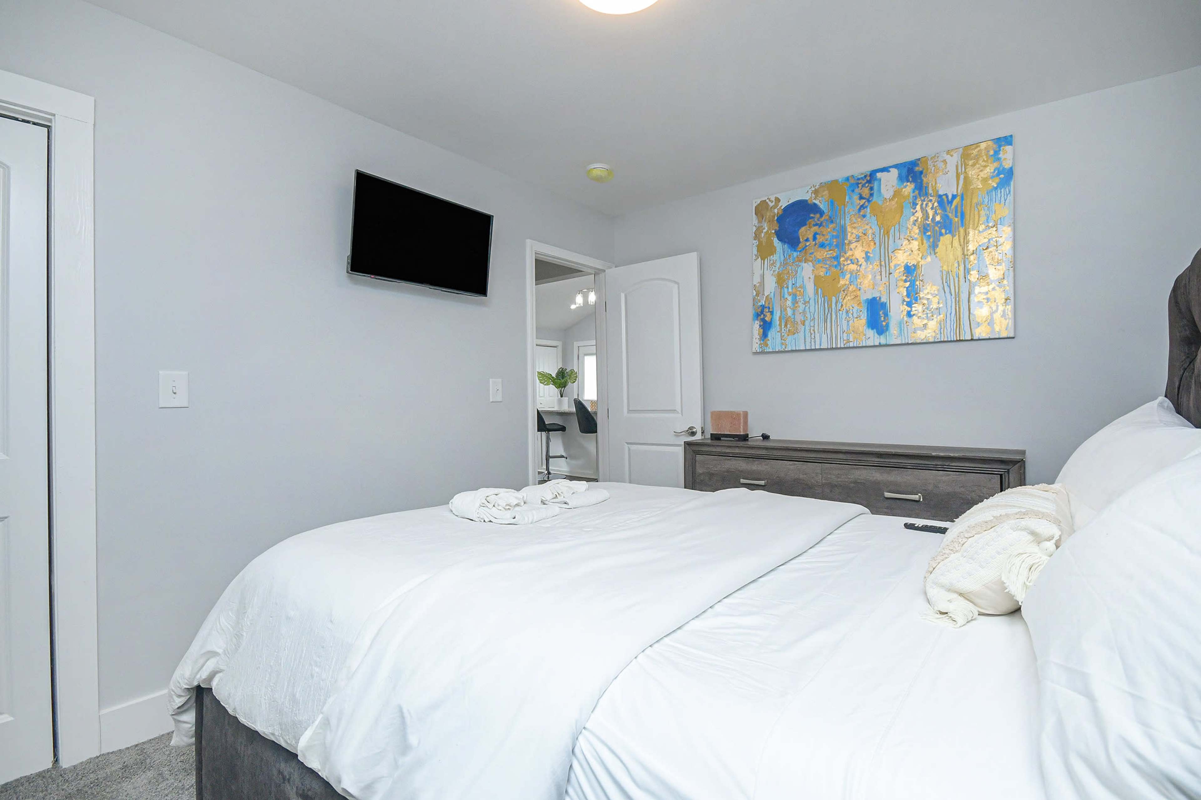 A neatly arranged bedroom with a bed, a television mounted on the wall, and a colorful piece of art above a dresser.