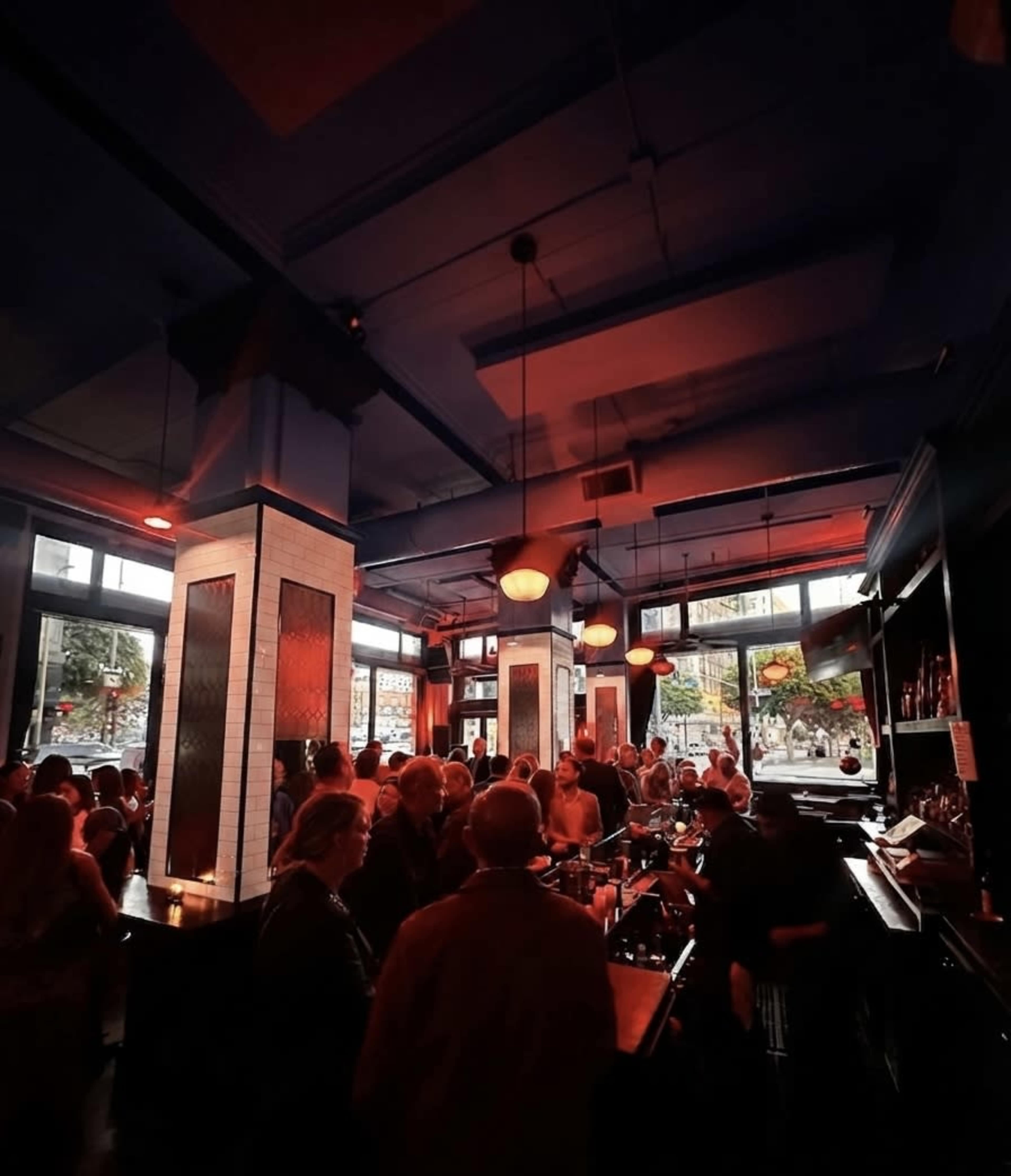 The image depicts a crowded bar interior with low red lighting and large windows allowing light to filter in.
