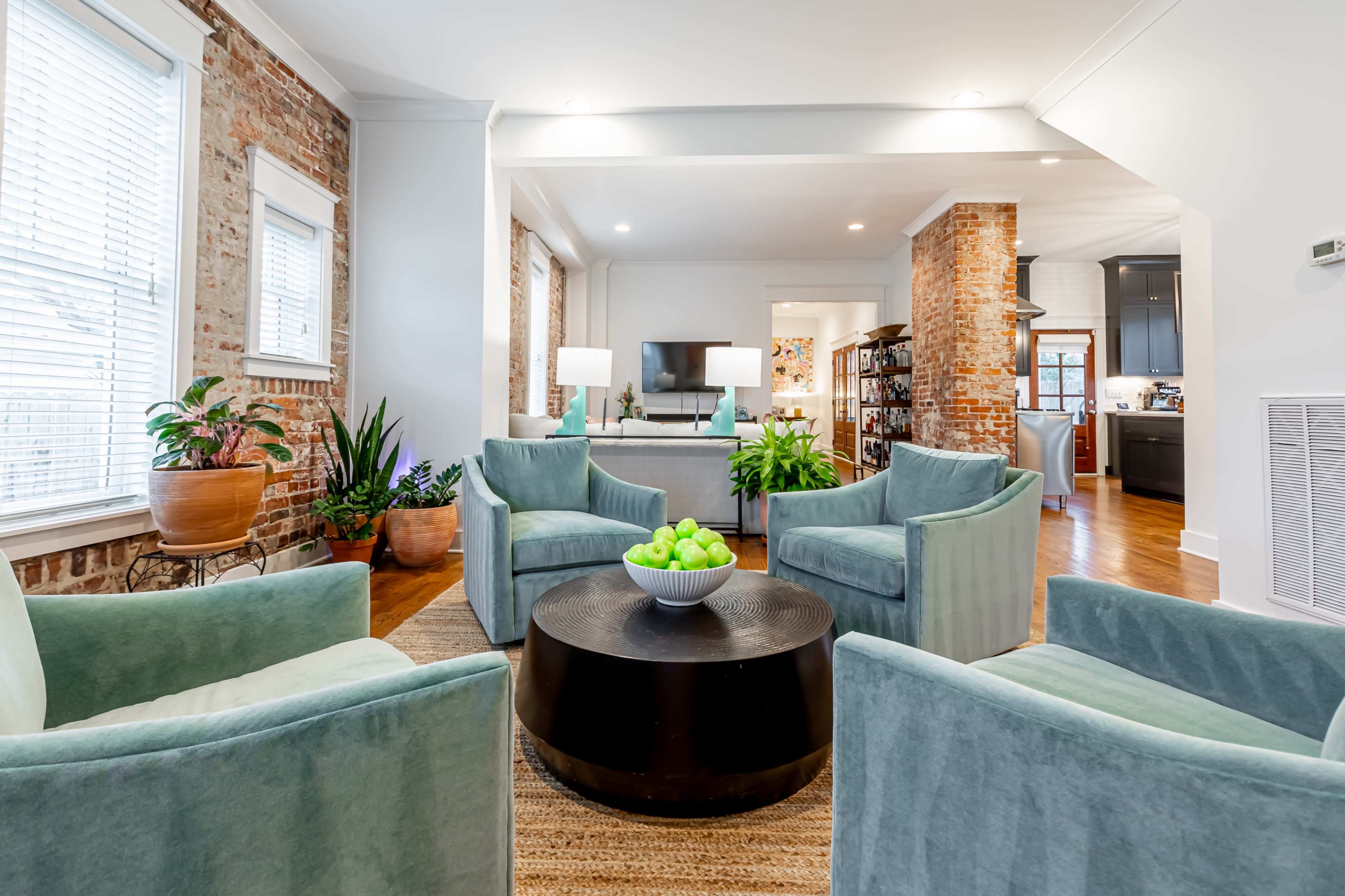 The image shows a modern living space featuring four upholstered chairs arranged around a round black table, with plants and a view into a kitchen area with exposed brick walls.